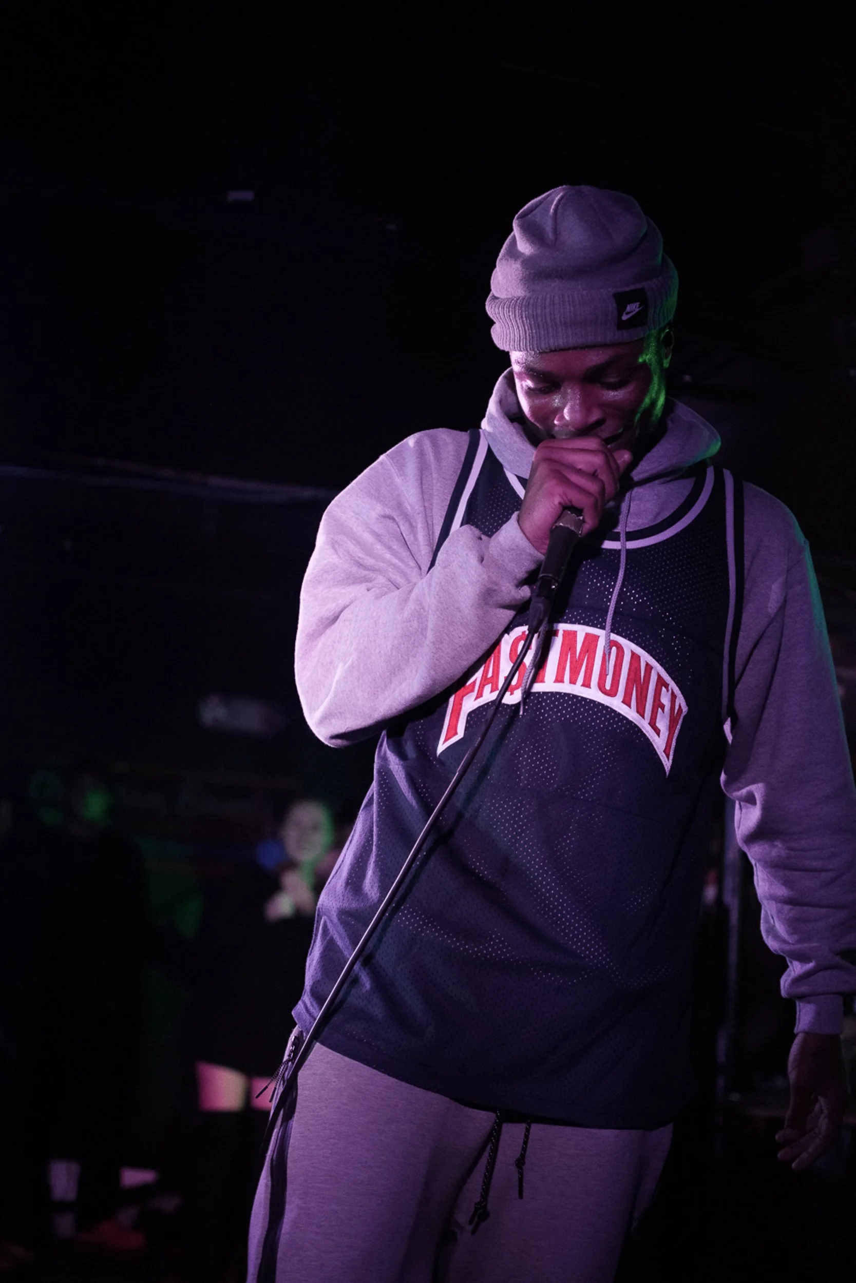 A person with a gray beanie and gray hoodie is holding a microphone and appears to be performing or speaking in a dark setting with purple and green lighting.