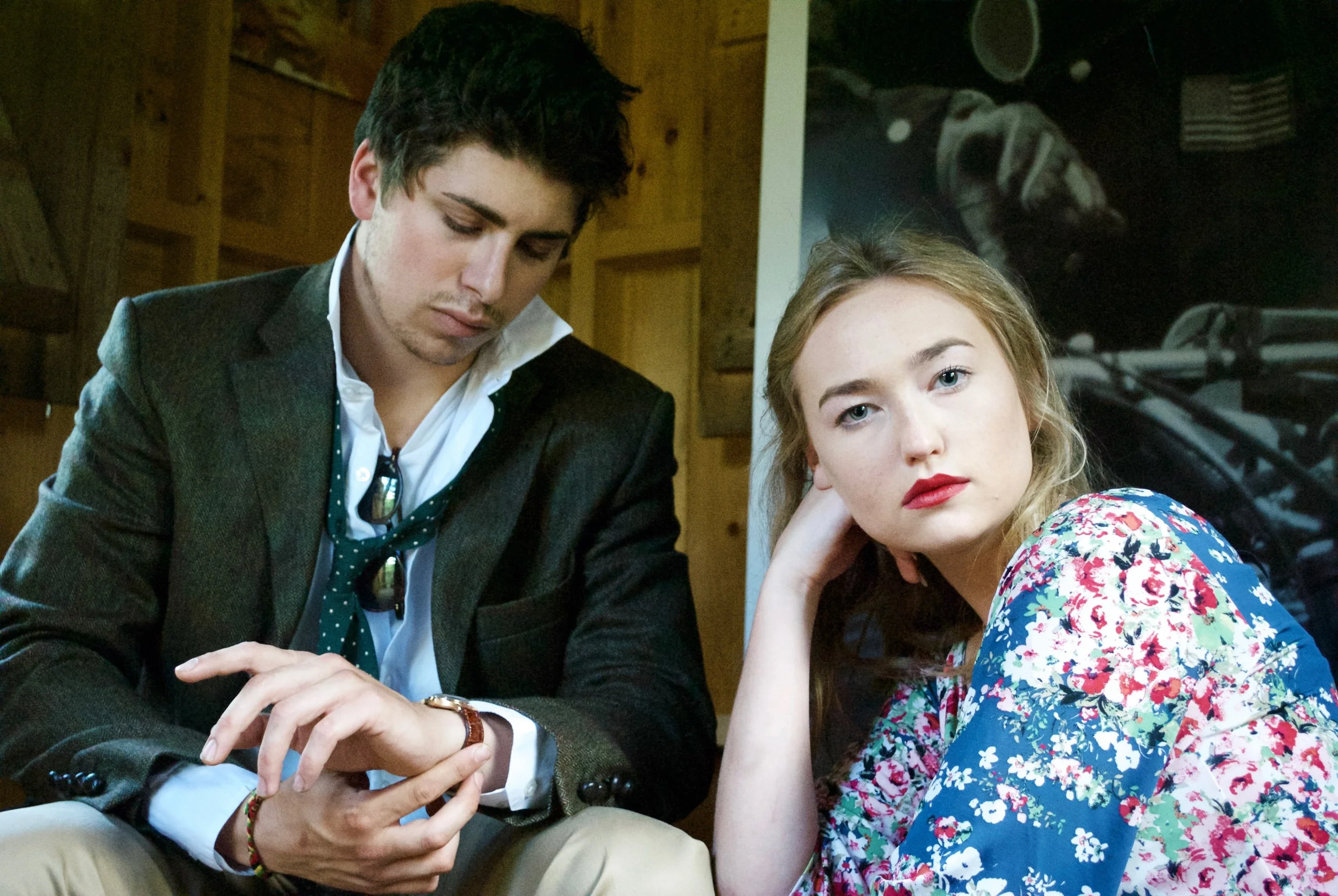 A man with dark hair in a blazer and white shirt looking down at a phone, and a woman with blonde hair and blue eyes wearing a colorful floral dress resting her head on her hand, sitting next to each other in an indoor setting.