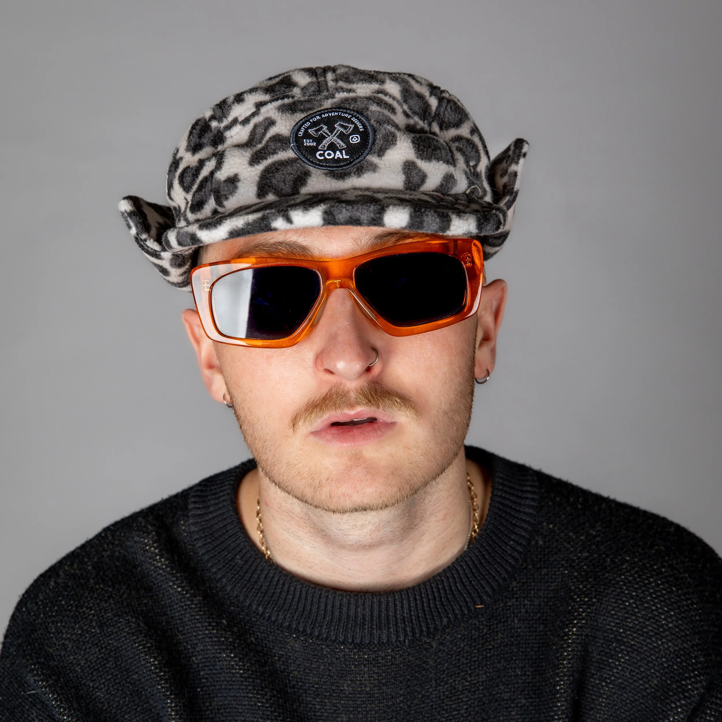Person wearing an animal print hat with a 'Coal' patch, orange sunglasses, and a black textured sweater, against a plain gray background.