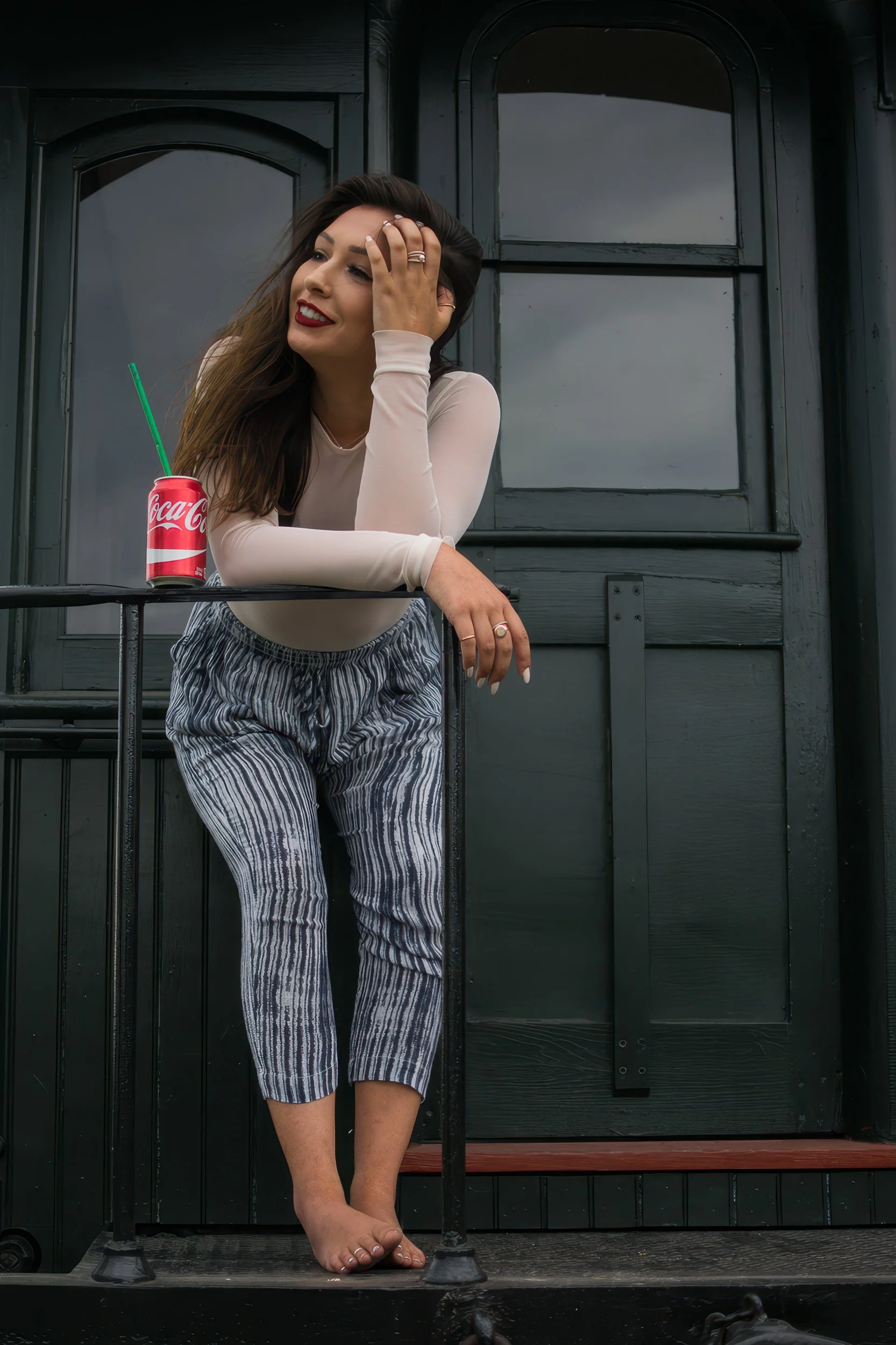 A young woman with long brown hair and red lipstick smiling, holding her forehead with one hand, and leaning on a black, metal railing of a small balcony. She is barefoot and wearing a long-sleeved cream top and striped trousers. There is a red Coca-
