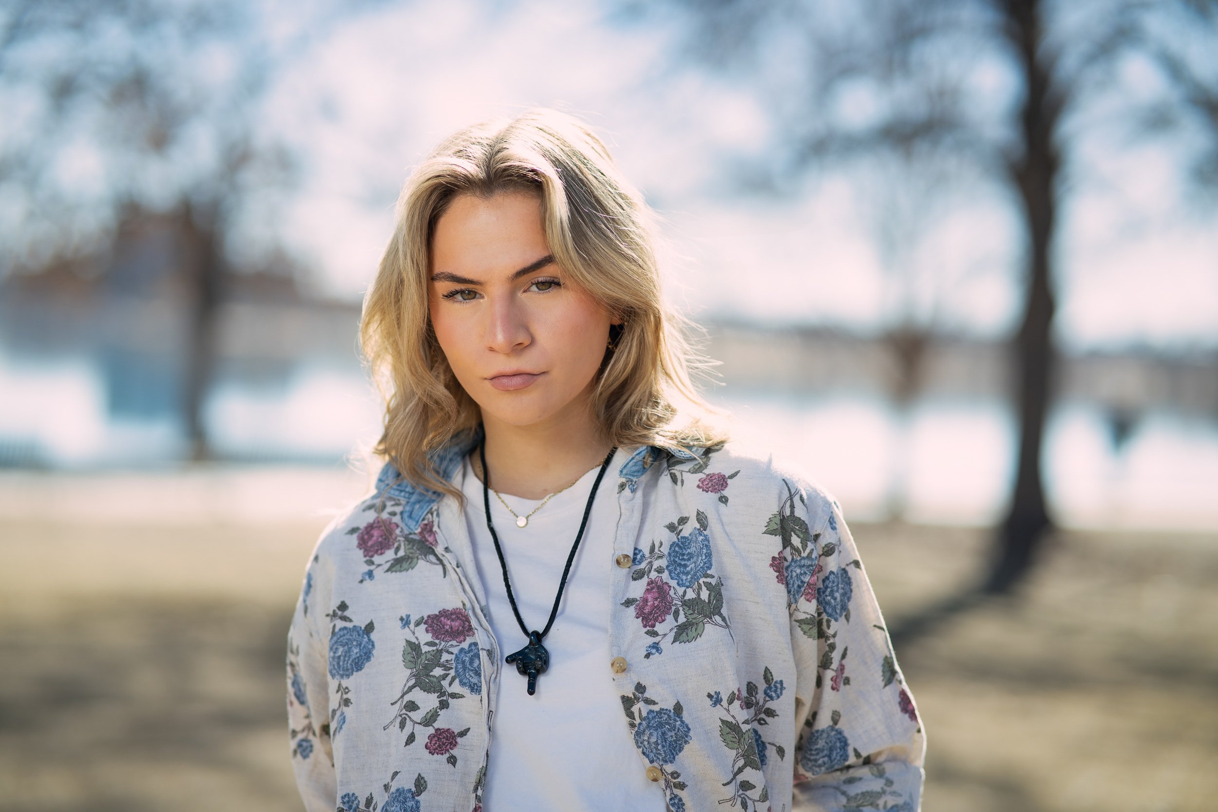 A young woman with blond hair and fair skin standing outdoors during daytime with a blurred background of trees and sky. She wears a floral jacket, a white shirt, and a necklace with a pendant, and looks directly at the camera.