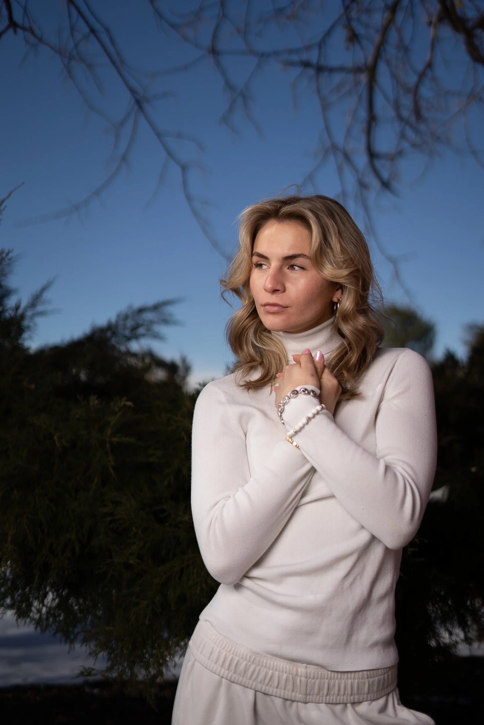 A young woman with blonde wavy hair wearing a white turtleneck sweater and white pants, standing outdoors at dusk or evening with trees and dark sky in the background.