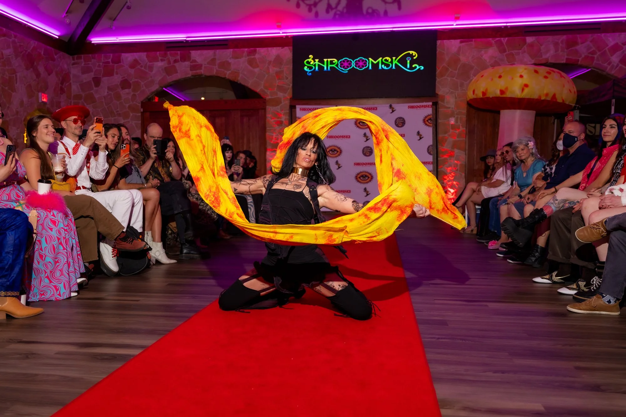 A dancer performing with yellow-orange fabric on a red carpeted runway in front of an audience and a backdrop with the word 'ShroomSky' in colorful neon lights. The audience is seated along the sides, taking photos and watching. The setting has brick