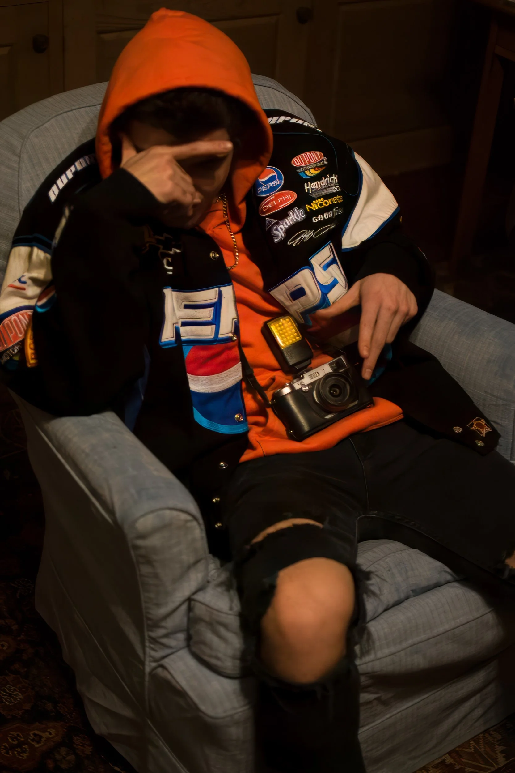 A person with a bald head is lying on a couch with their head turned downward, wearing a gray shirt with ripped sleeve. Another person, wearing a jacket with various patches and a red hood, is sitting next to them, covering part of their face with on