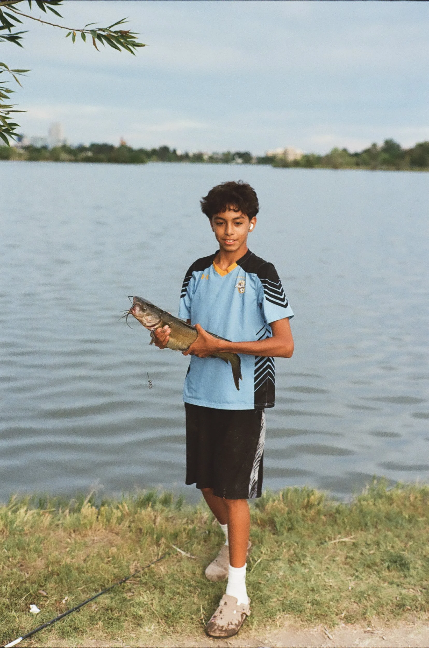 A boy wearing a light blue sports shirt and black shorts with African American features standing by a lake holding a fish in his hands. There are trees and buildings in the background, and the boy is standing on grass with a fishing rod on the ground