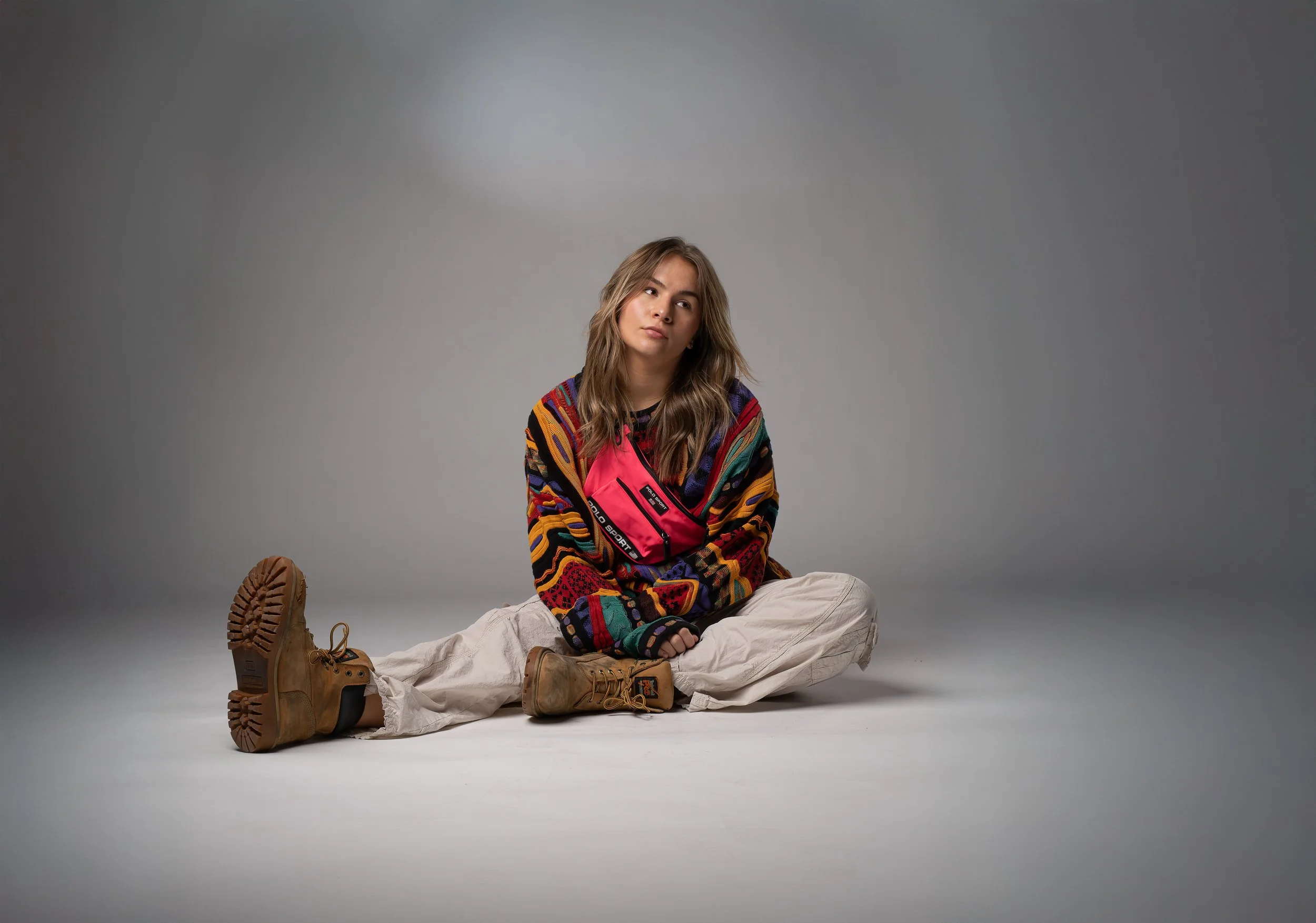 A young woman with long wavy hair sitting on the floor against a plain gray background. She is wearing a colorful, patterned sweater, beige pants, and tan combat boots. She appears thoughtful or contemplative.