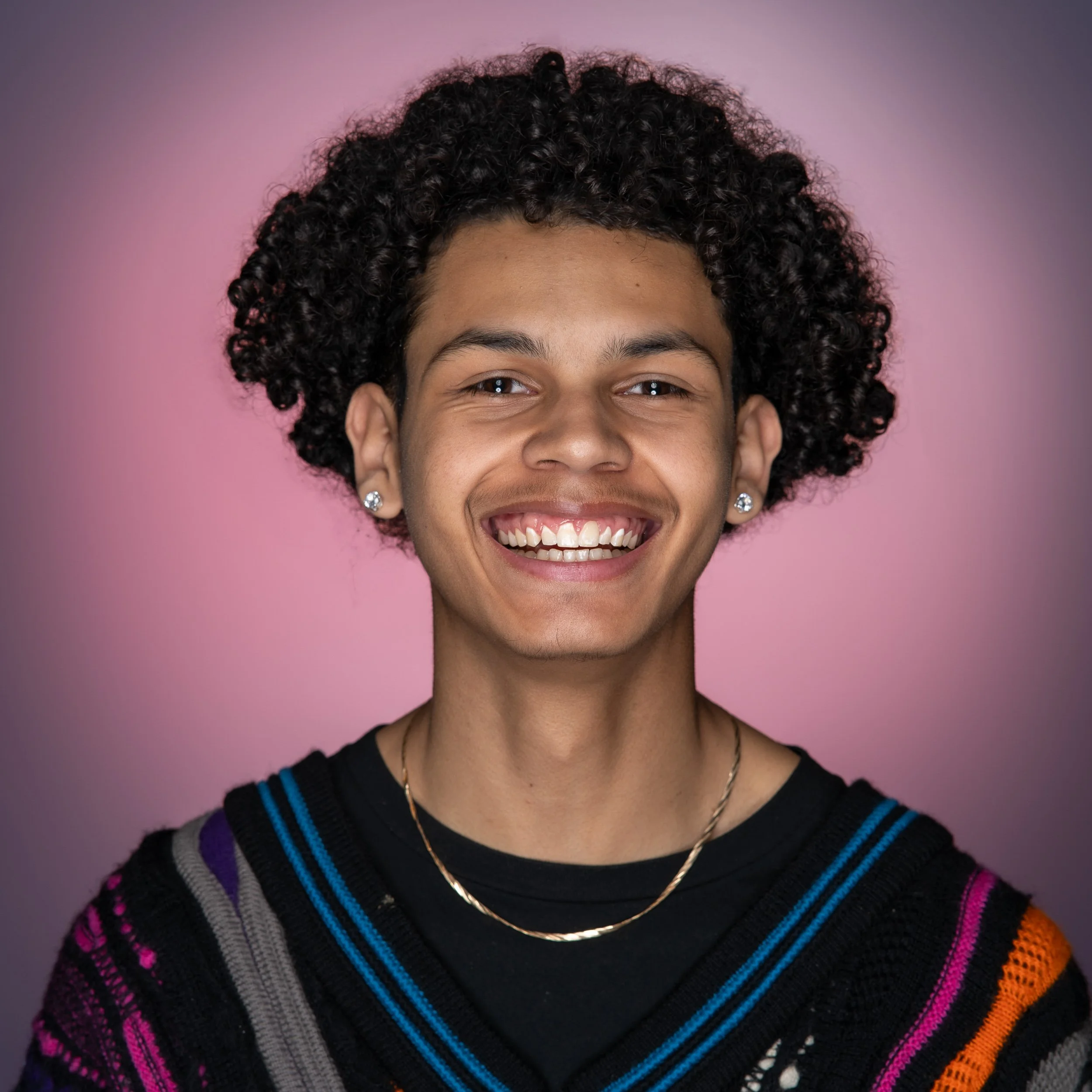 Close-up of a person's face with curly hair, smiling, wearing a colorful rainbow sweater and jewelry, against a pink background.