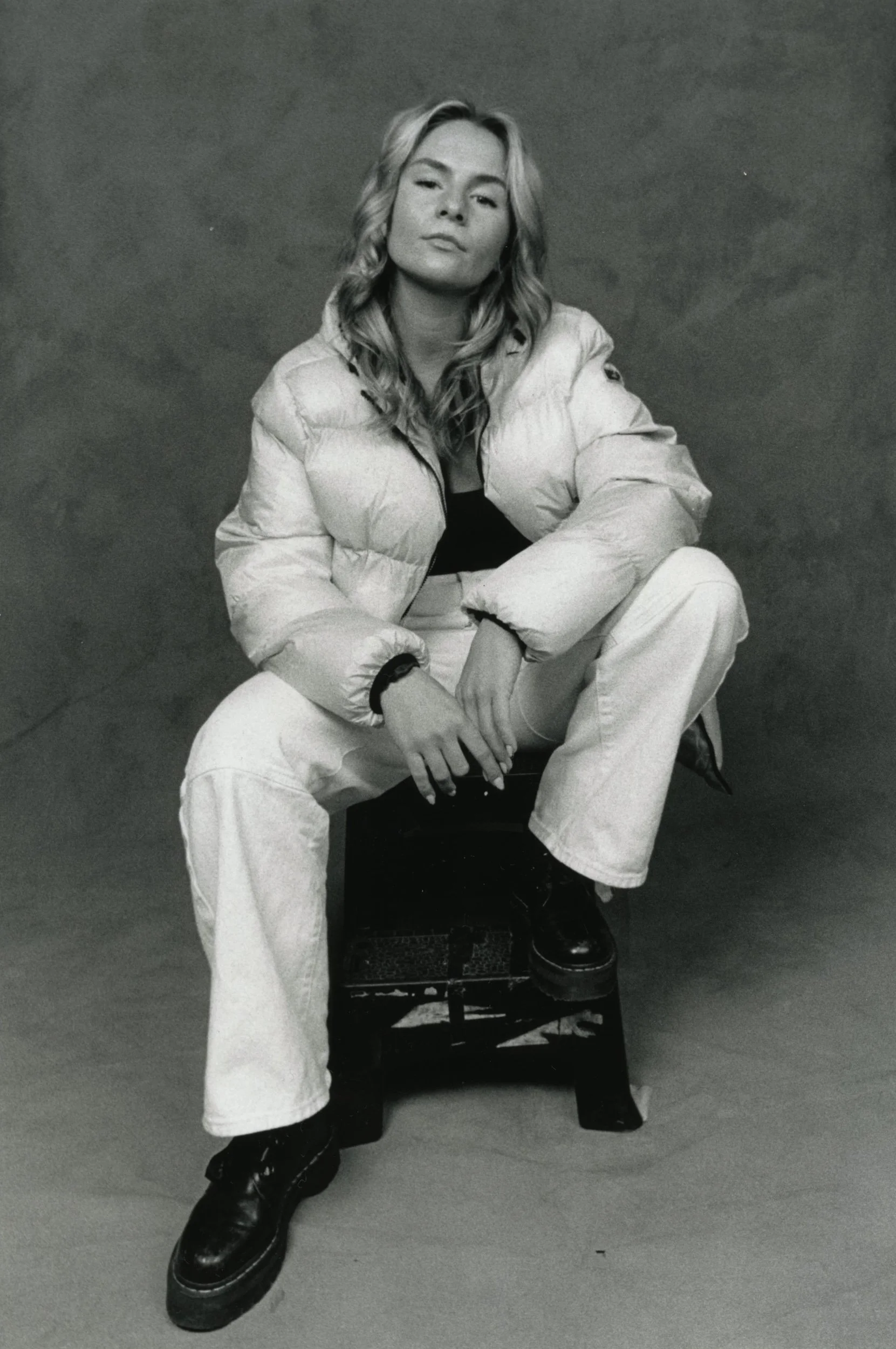 Black and white photo of a woman with wavy hair, wearing a puffy jacket, sitting on a chair with one leg crossed over the other, against a plain background.