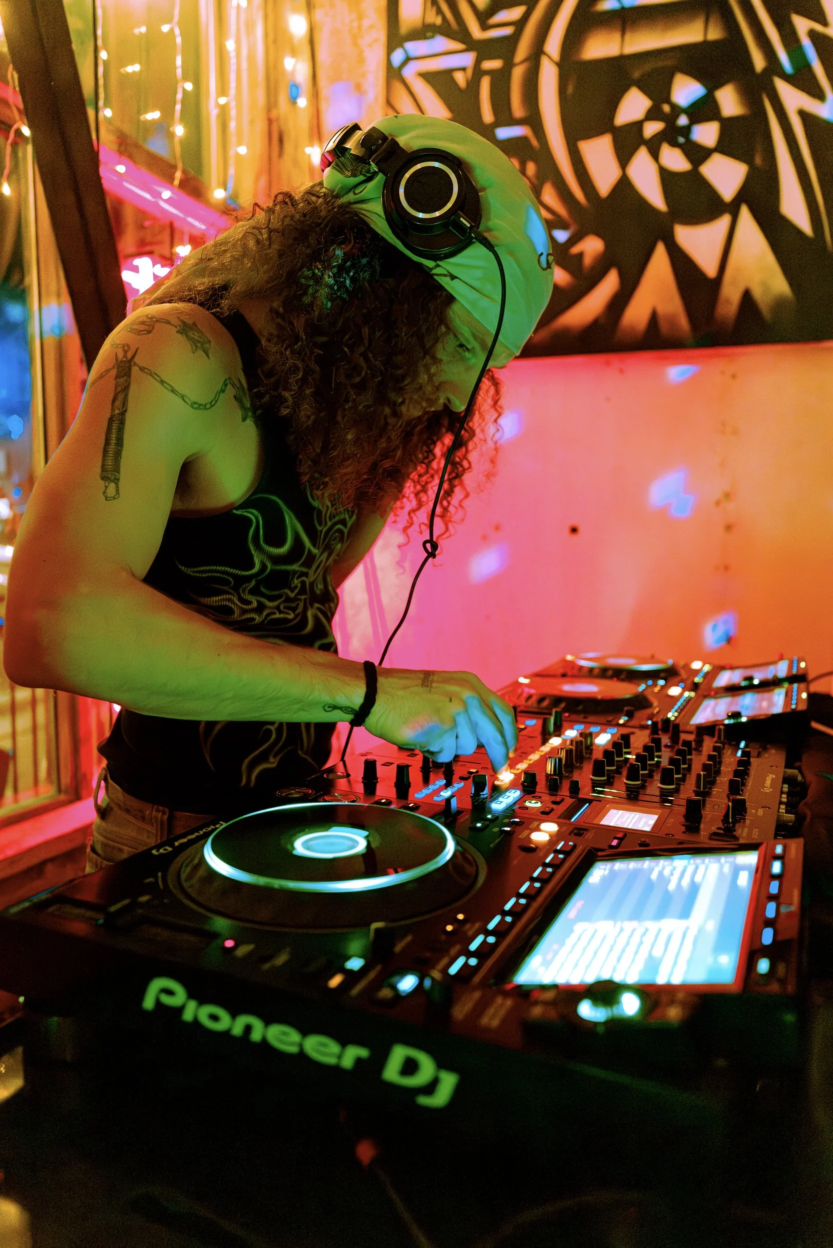 A DJ wearing a yellow bandana, headphones, and a black tank top, mixing music at a vibrant, colorful party. The background is decorated with bright lights and abstract patterns.