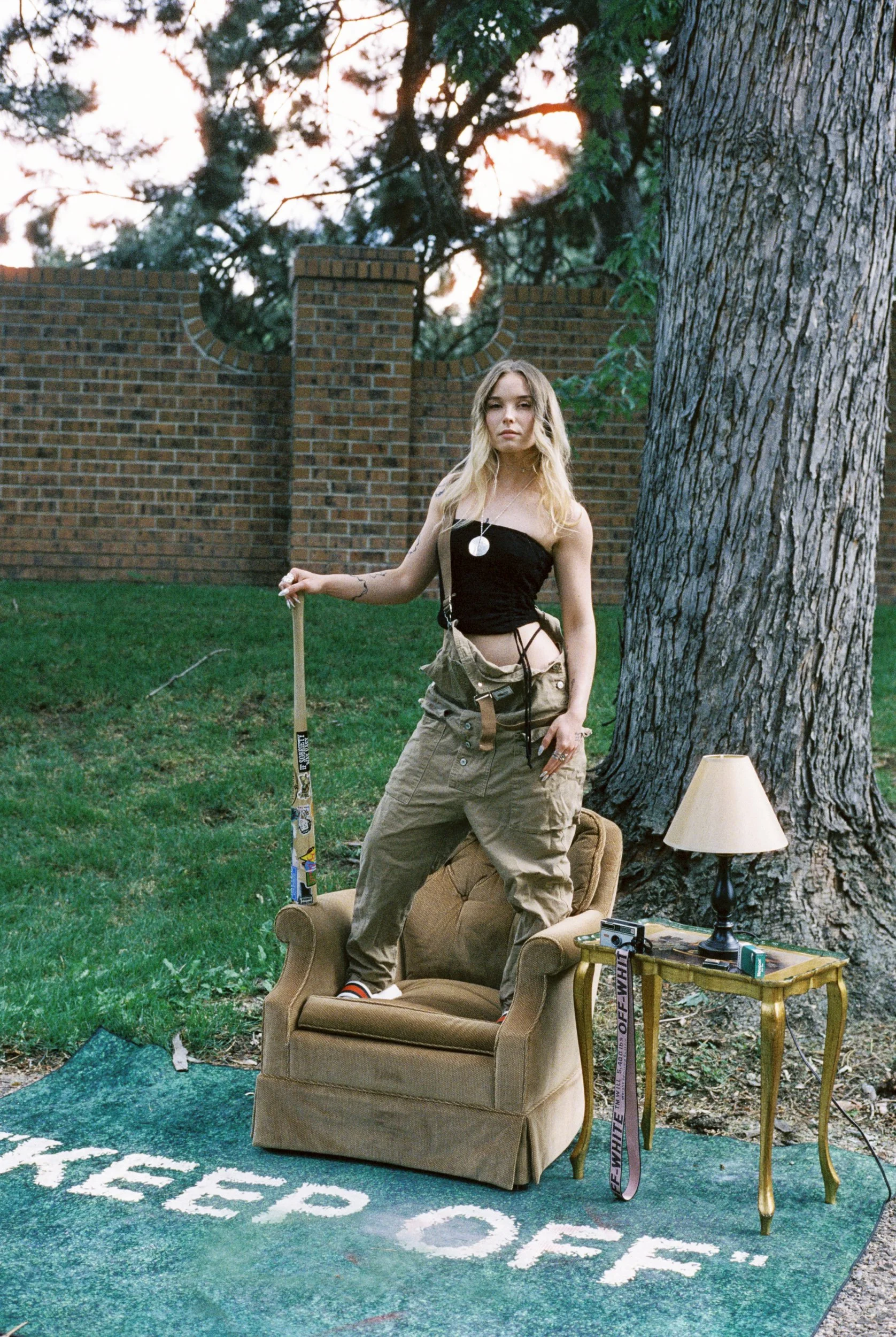 A young woman with blonde hair standing outdoors next to a large tree, posed with one leg resting on an armchair inside a decorative outdoor setting, with a brick wall and greenery in the background, and various items including a lamp, backpack, and 