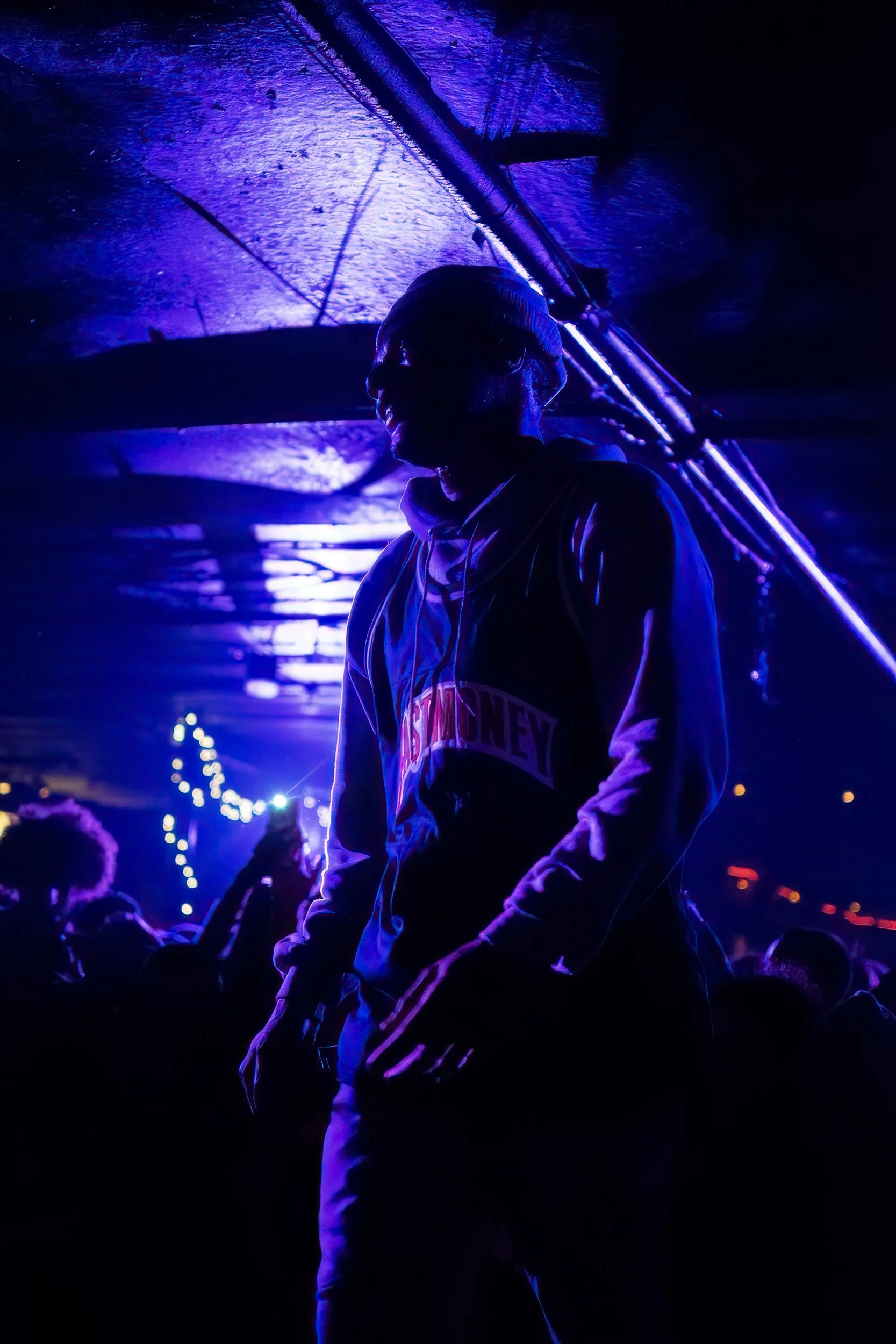 Person in a hoodie and glasses standing in a crowd at night illuminated by purple and blue neon lights.