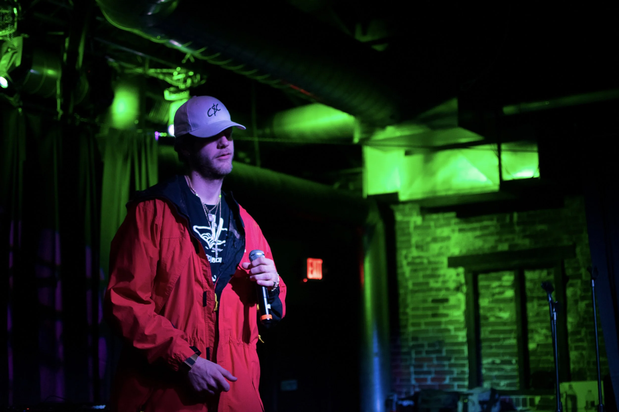 A person wearing a white cap, red jacket, and black shirt standing in a dimly lit room with green and purple lighting, holding a microphone.