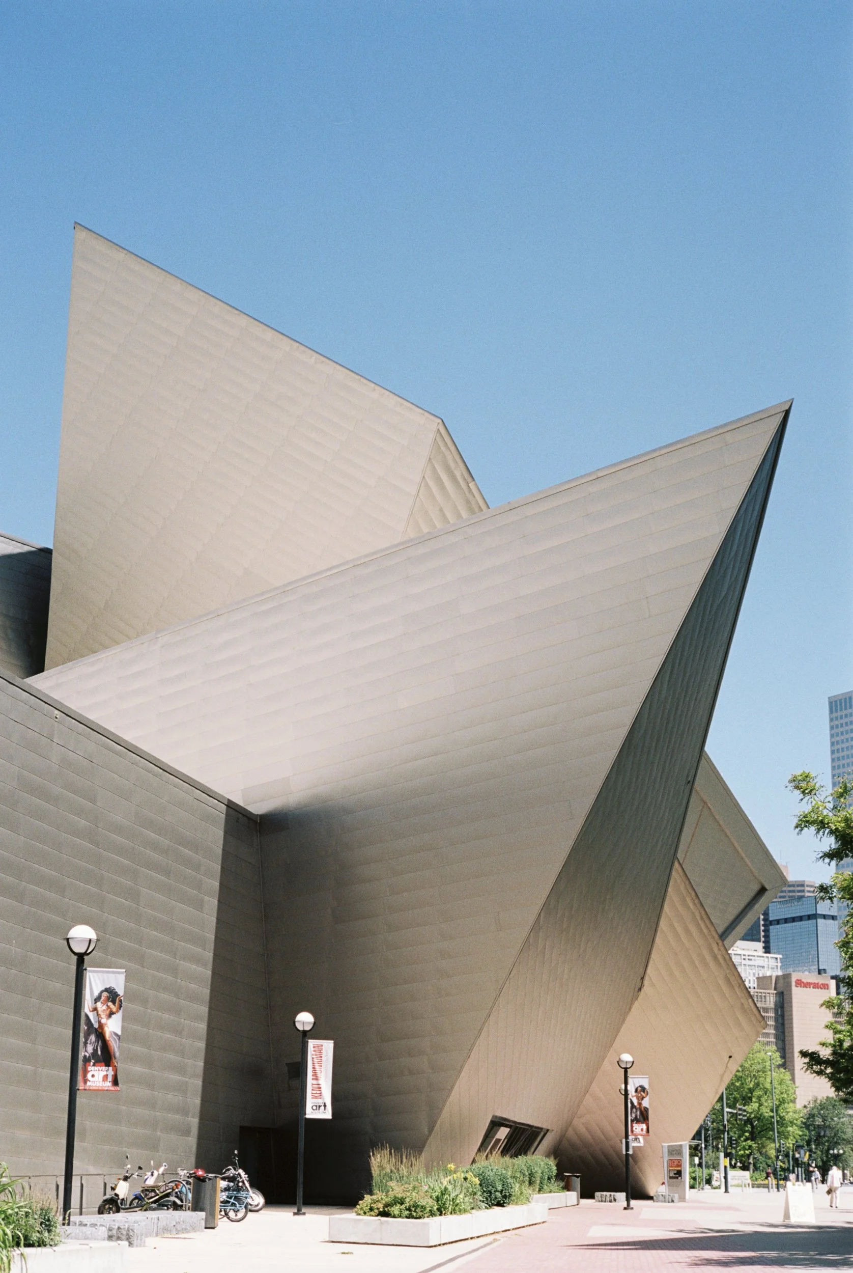 Modern building with angular, silver-colored metallic panels under a clear blue sky, with lampposts, banners, bikes, and greenery in the foreground.