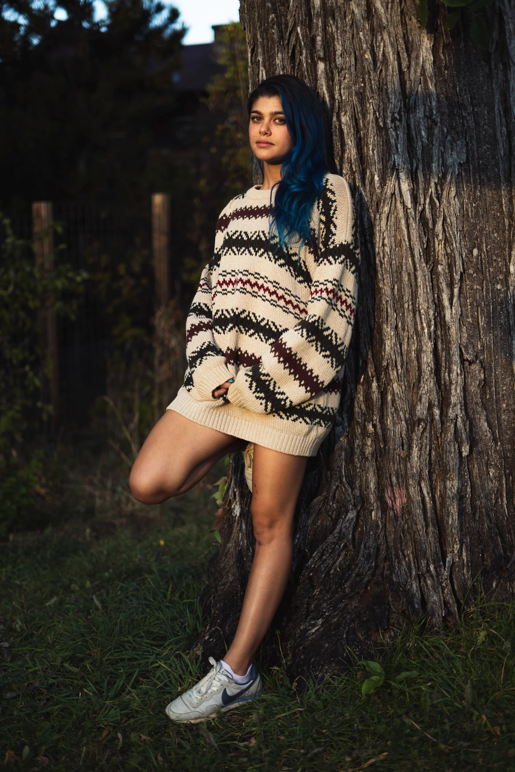 Young woman with blue hair leaning against a large tree in a grassy outdoor area during late afternoon or early evening.