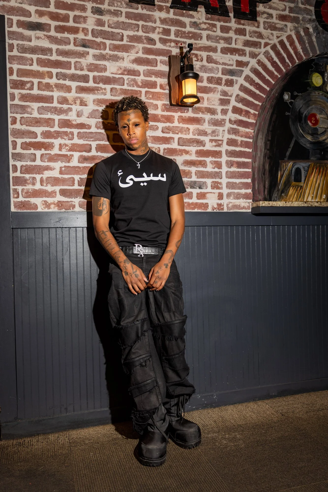A young man with tattoos standing against a brick wall with a black wood panel at the bottom. He wears a black T-shirt with white Arabic script, black cargo pants, and black boots, and has a serious expression.