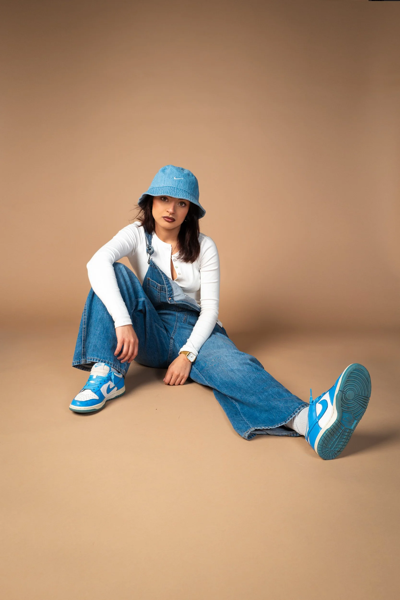 A woman sitting on the floor against a plain brown background. She is wearing a light blue bucket hat, a white long-sleeve shirt, blue jeans, and blue and white sneakers. She has dark hair and is looking at the camera with a neutral expression.
