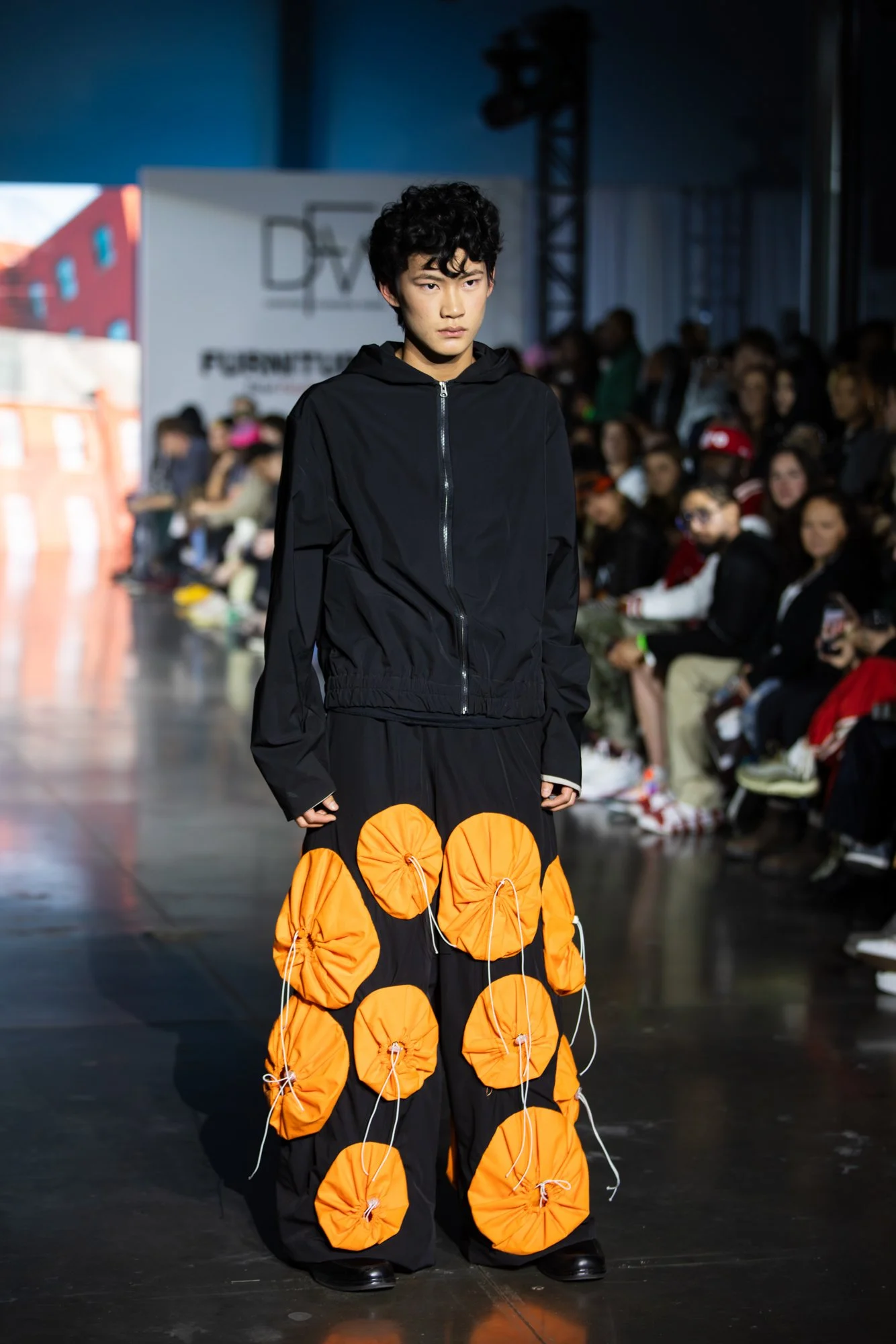 A male model walking on a runway during a fashion show, wearing a black hoodie and wide black pants with orange circular fabric pieces attached with drawstrings, in front of an audience with some standing and some seated, with a sign that reads "DW" 