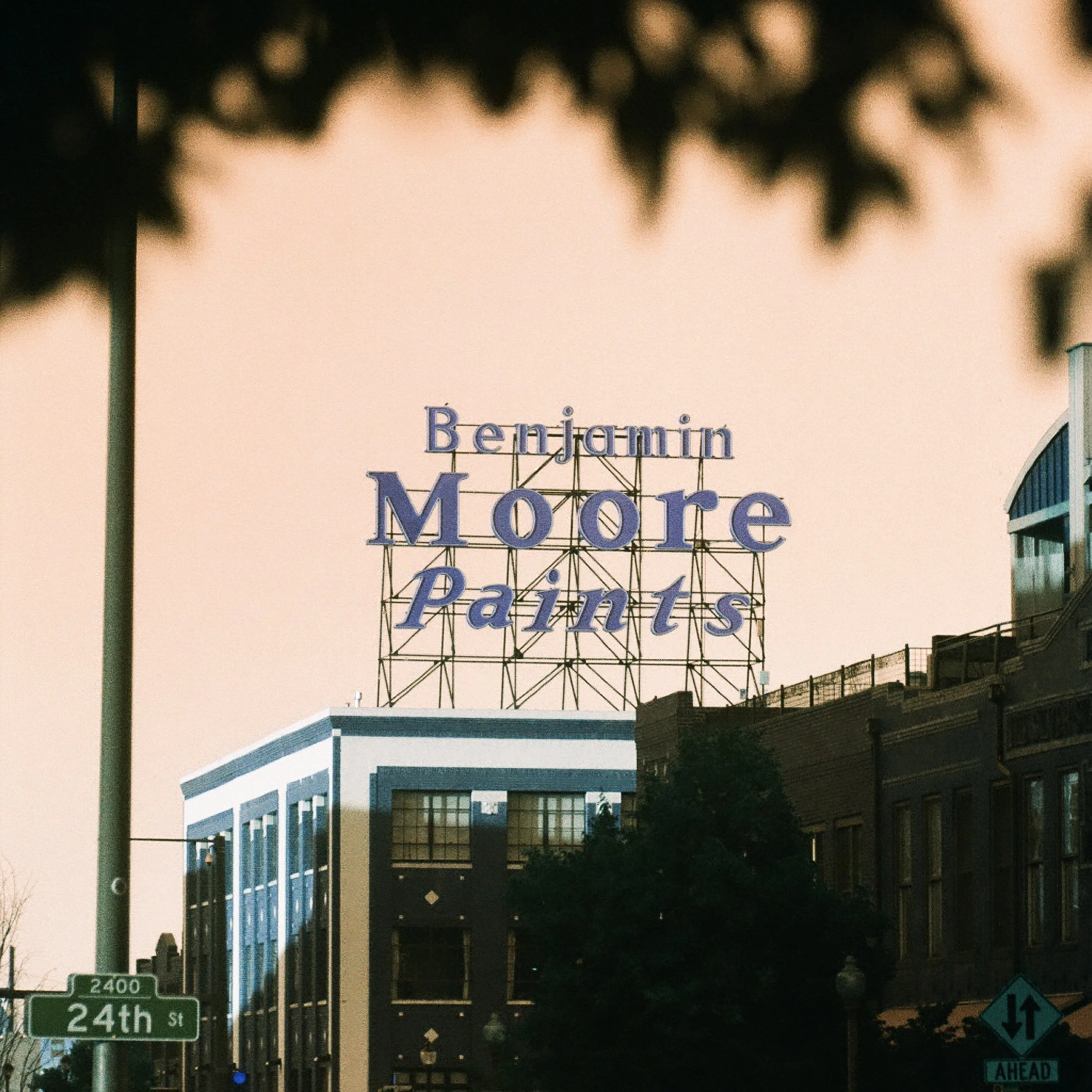Nighttime street scene with building signs, including a prominent sign for 'Benjamin Moore Paints', traffic signs, and street name signs for 24th Street and 2400 block.