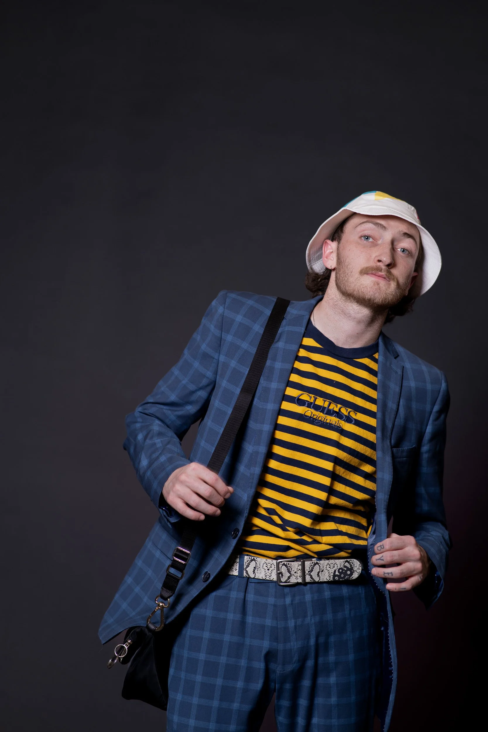 A man with shoulder-length brown hair and a beard, wearing a white bucket hat, a blue plaid blazer, a yellow and black striped 'GUESS Originals' t-shirt, patterned belt, and carrying a black bag with a strap, posing against a dark plain background.