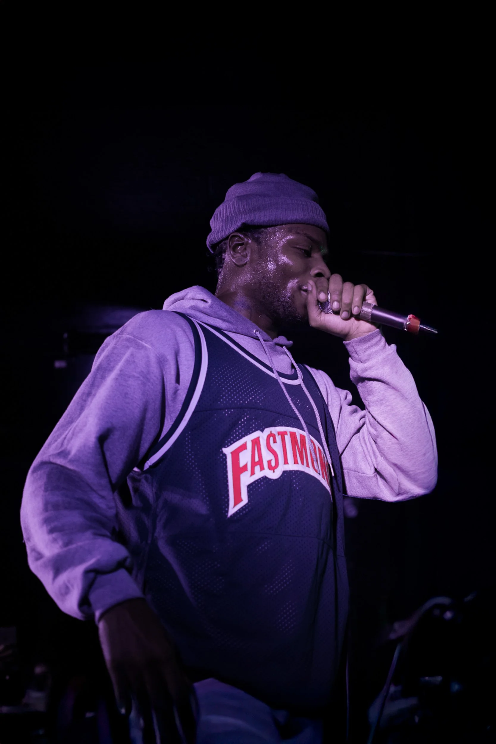 A young man wearing a purple beanie and hoodie, holding a microphone close to his mouth, appears to be performing on stage under purple lighting, with sweat visible on his face.