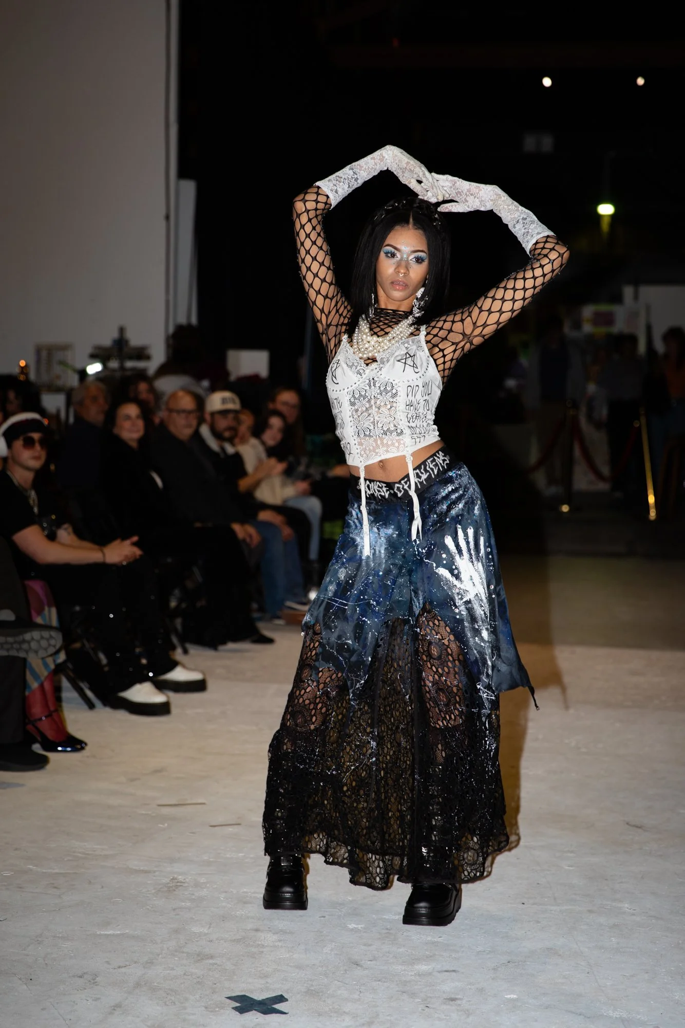 Model walking down runway in black lace and mesh dress layered over blue galaxy-print skirt, wearing white gloves and layered pearl necklaces, with audience watching.