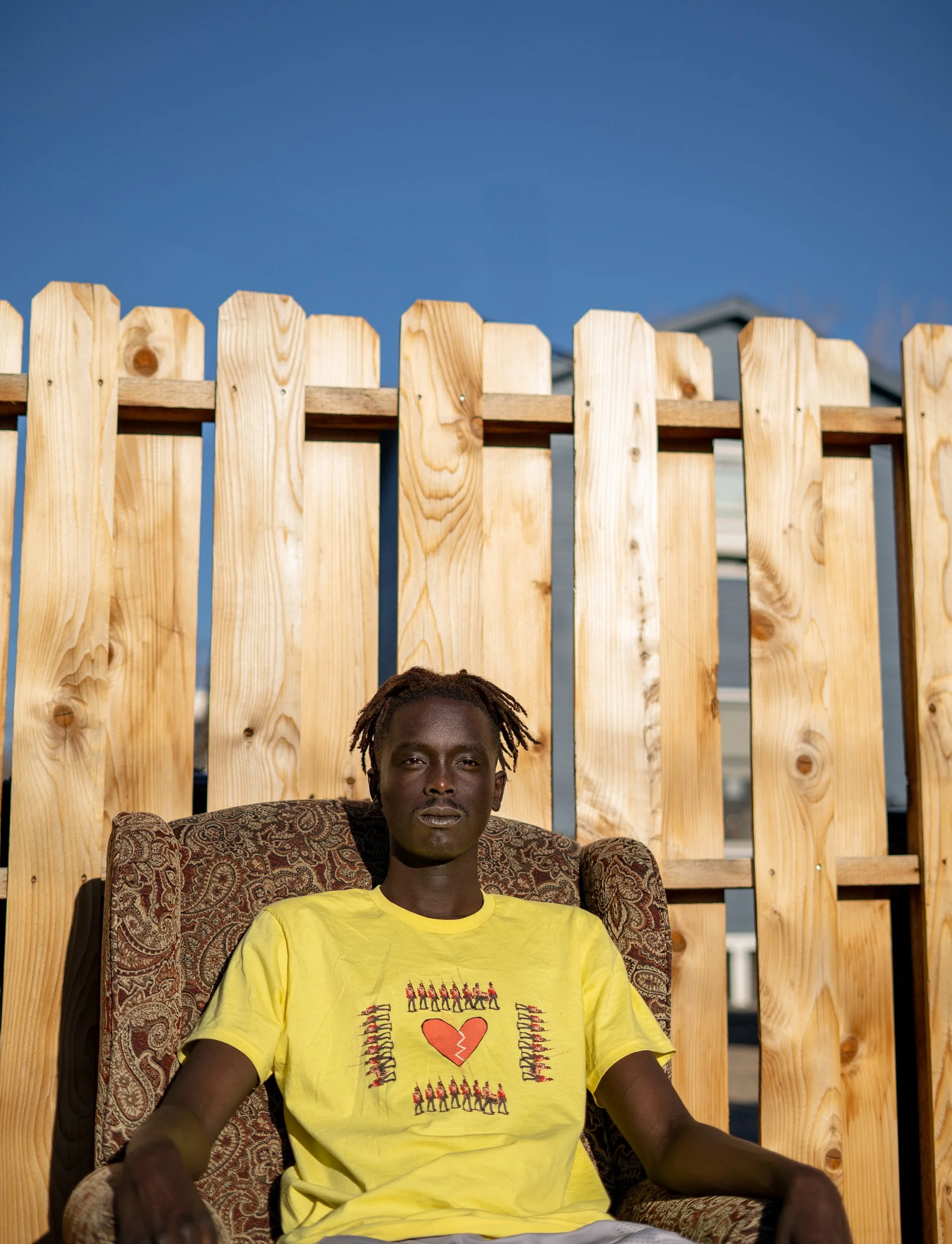 A young man with dark skin and dreadlocks sitting in a patterned armchair in front of a tall wooden fence, wearing a yellow T-shirt with a heart and soldier illustrations.