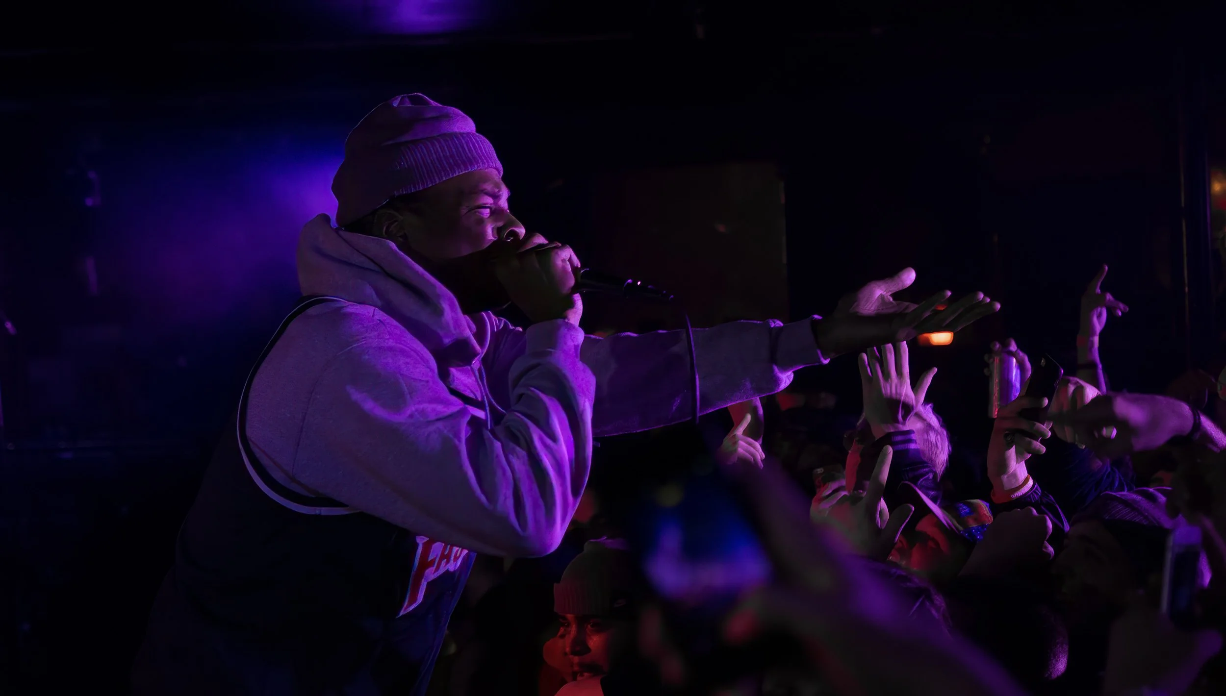 Performer wearing a purple beanie and hoodie singing into a microphone during a concert, surrounded by audience members with hands raised