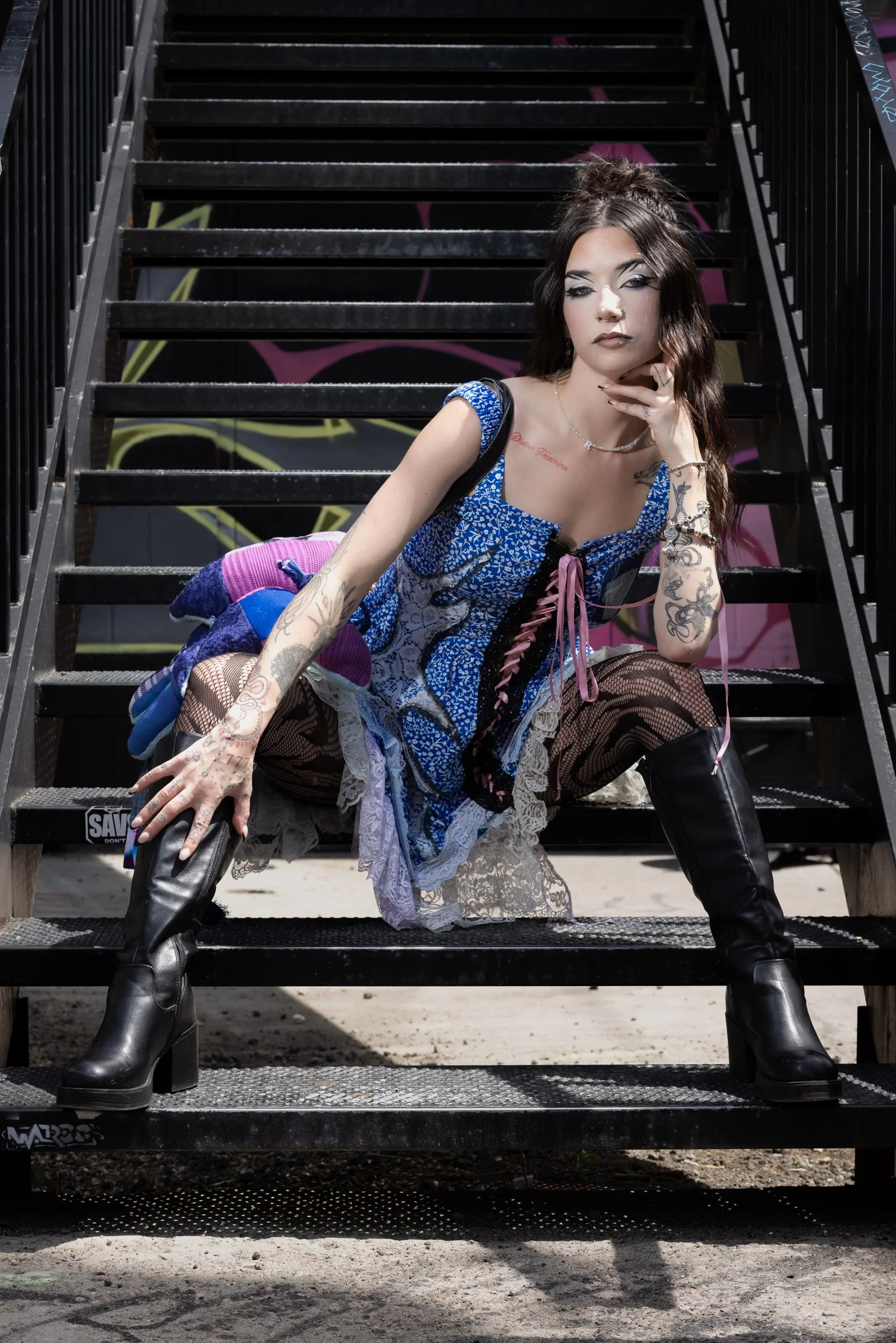 A woman with long dark hair and tattoos on her arms and chest, wearing a blue patterned dress, fishnet tights, and black boots, sits on metal stairs with graffiti background, striking a pose with her hand near her face.