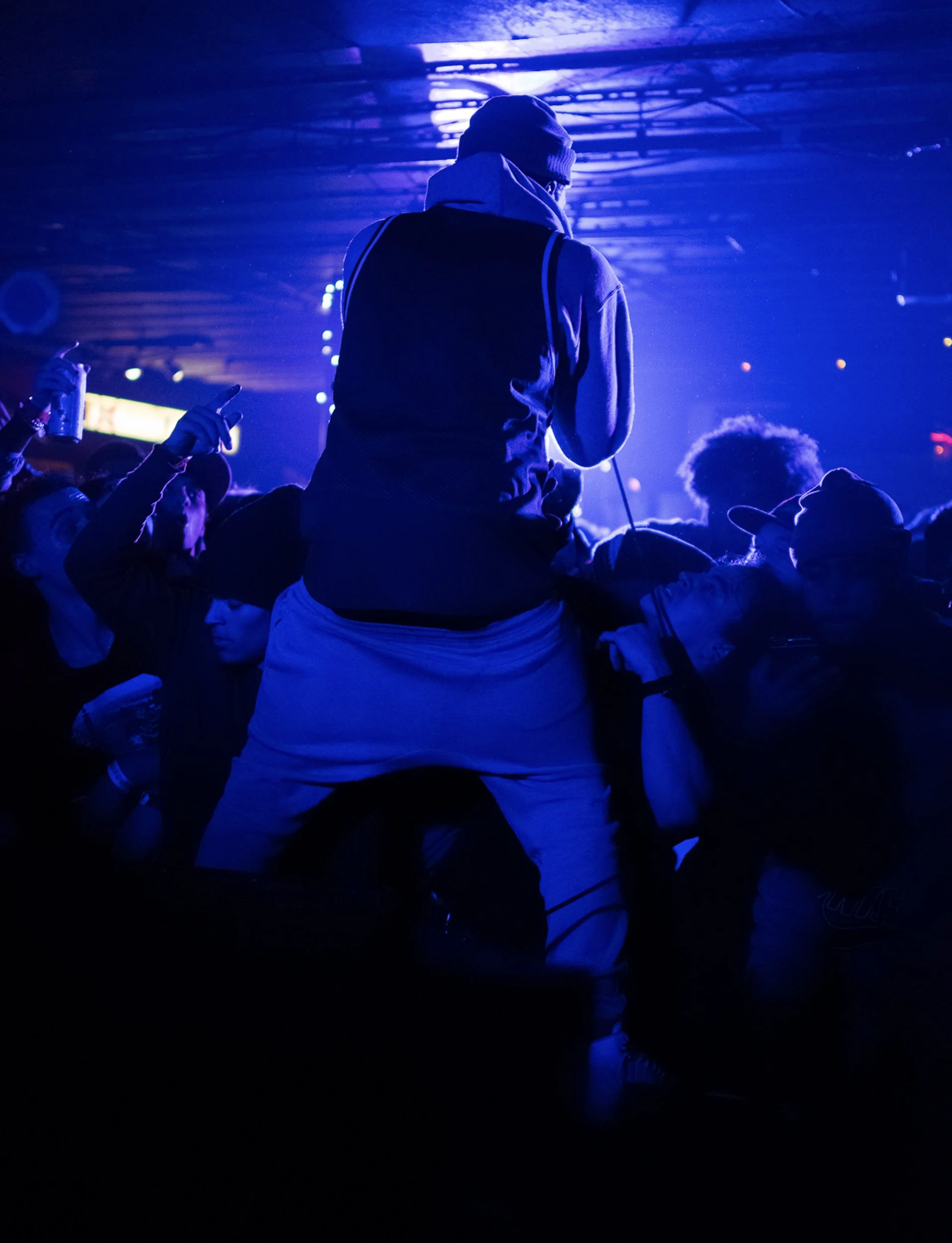 A person standing with their back to the camera at a concert or nightclub, illuminated by blue and purple lights, with a crowd around them.