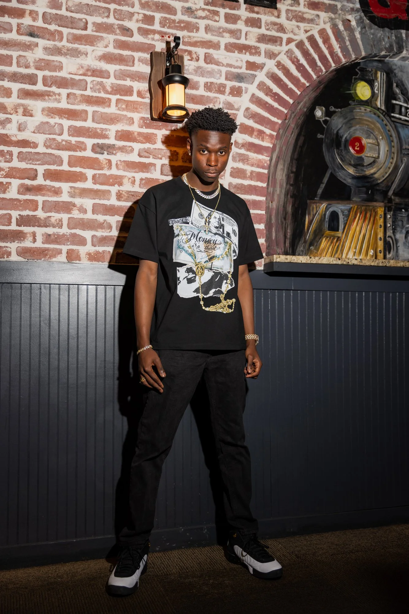 A young man standing against a brick wall with a black and gray paneling below, wearing a graphic t-shirt, black pants, sneakers, and jewelry, with a brick wall and a large mechanical object in the background.