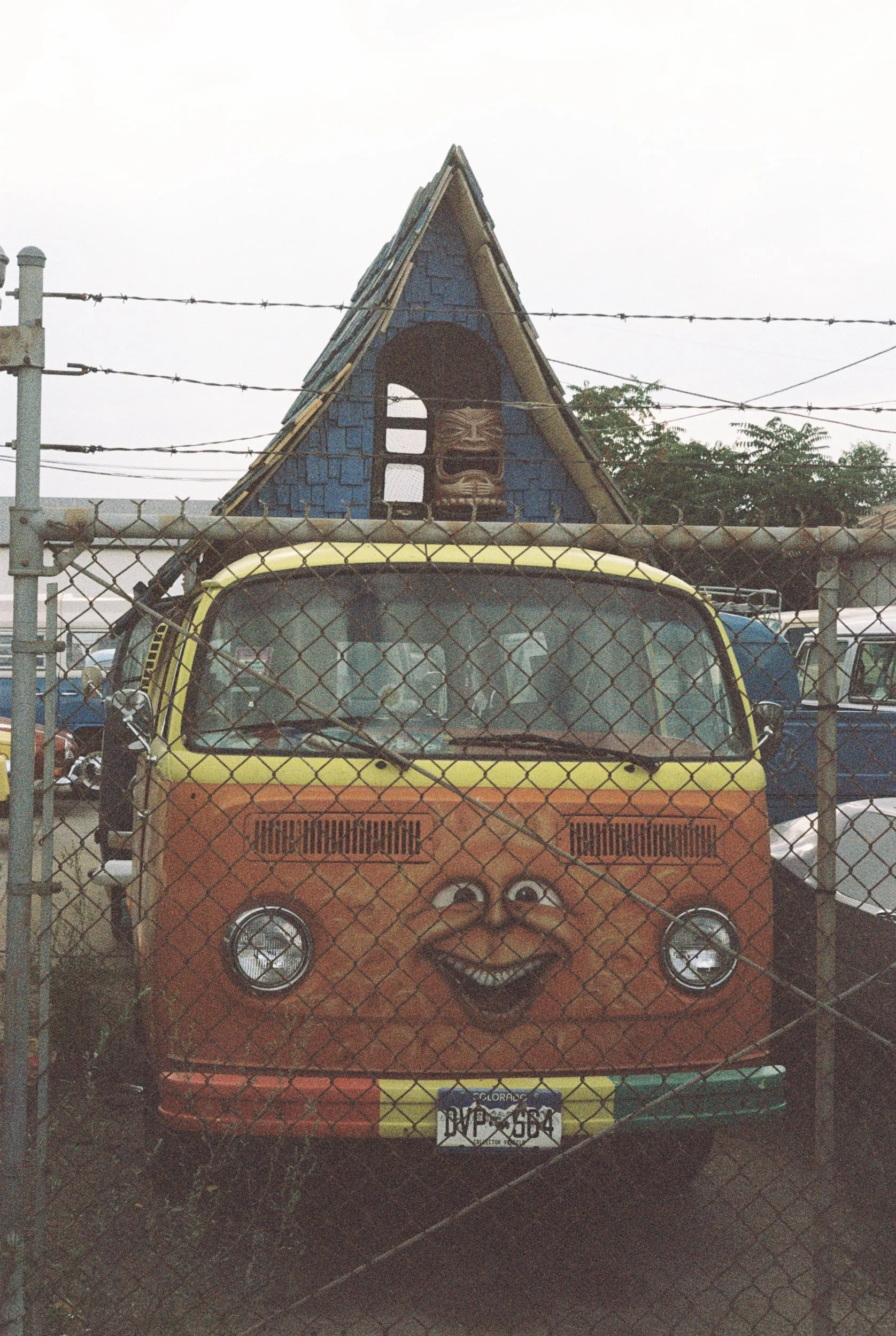 A truck decorated with a cartoon face, including big eyes and a wide smile, is parked behind a chain-link fence. The background shows an A-frame building with a tiki face cutout in the upper window and barbed wire overhead.