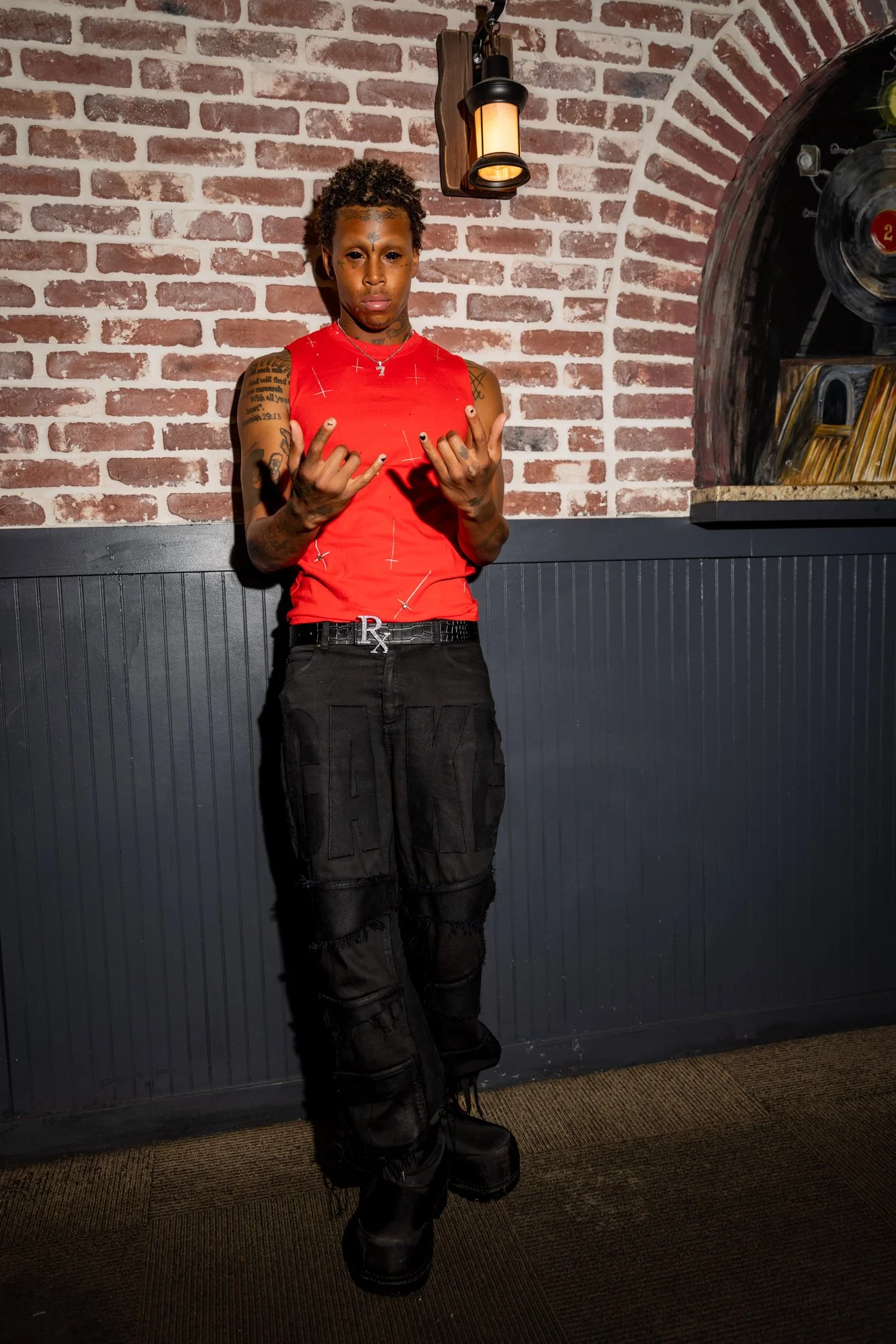 A young person with tattoos standing in front of a brick wall, wearing a red sleeveless top, black cargo pants, and black boots, making a hand gesture with both hands.