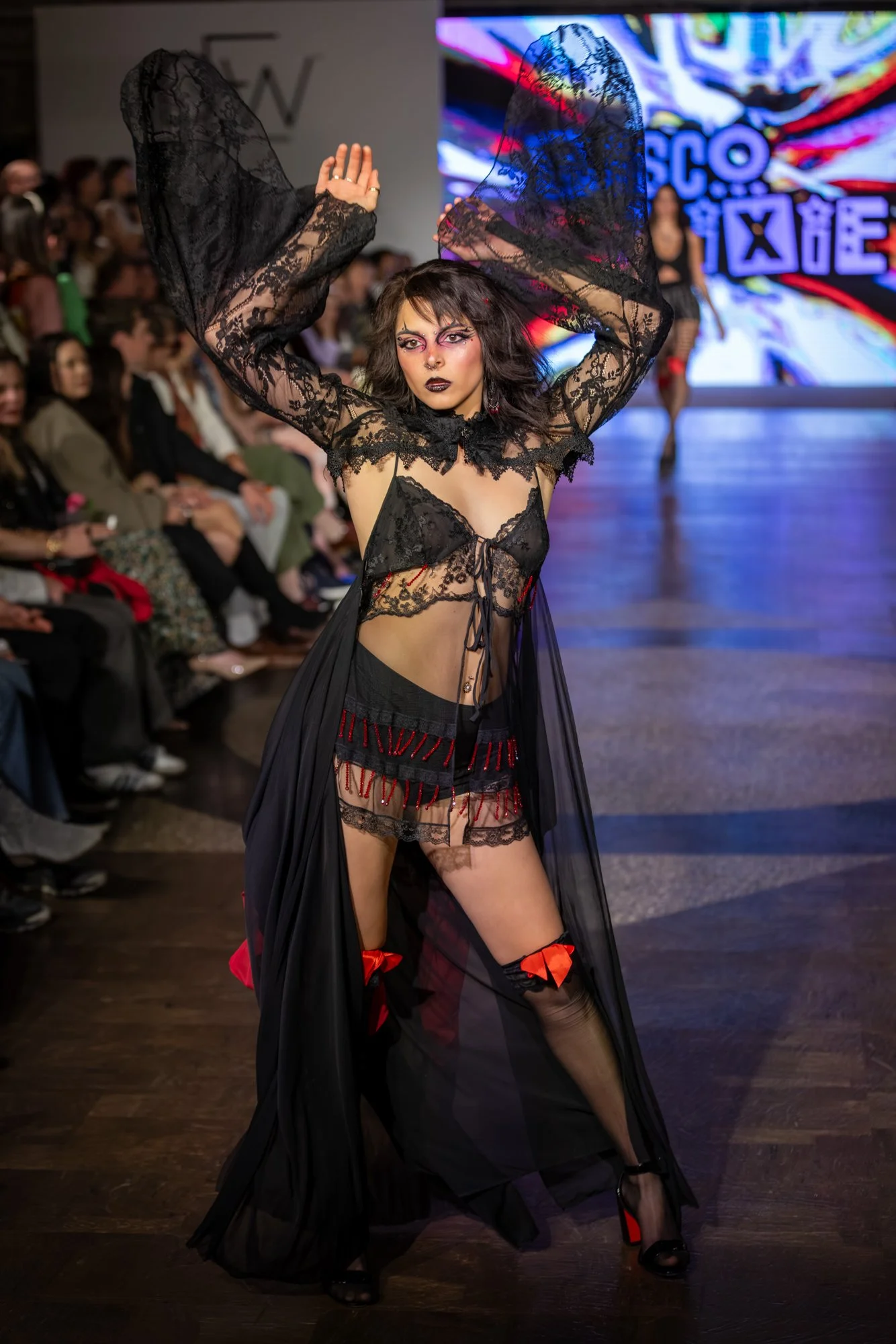 A fashion model walking on runway wearing black lace lingerie with sheer cape, thigh-high stockings with red bows, and dramatic makeup, audience watching in background and a colorful LED screen