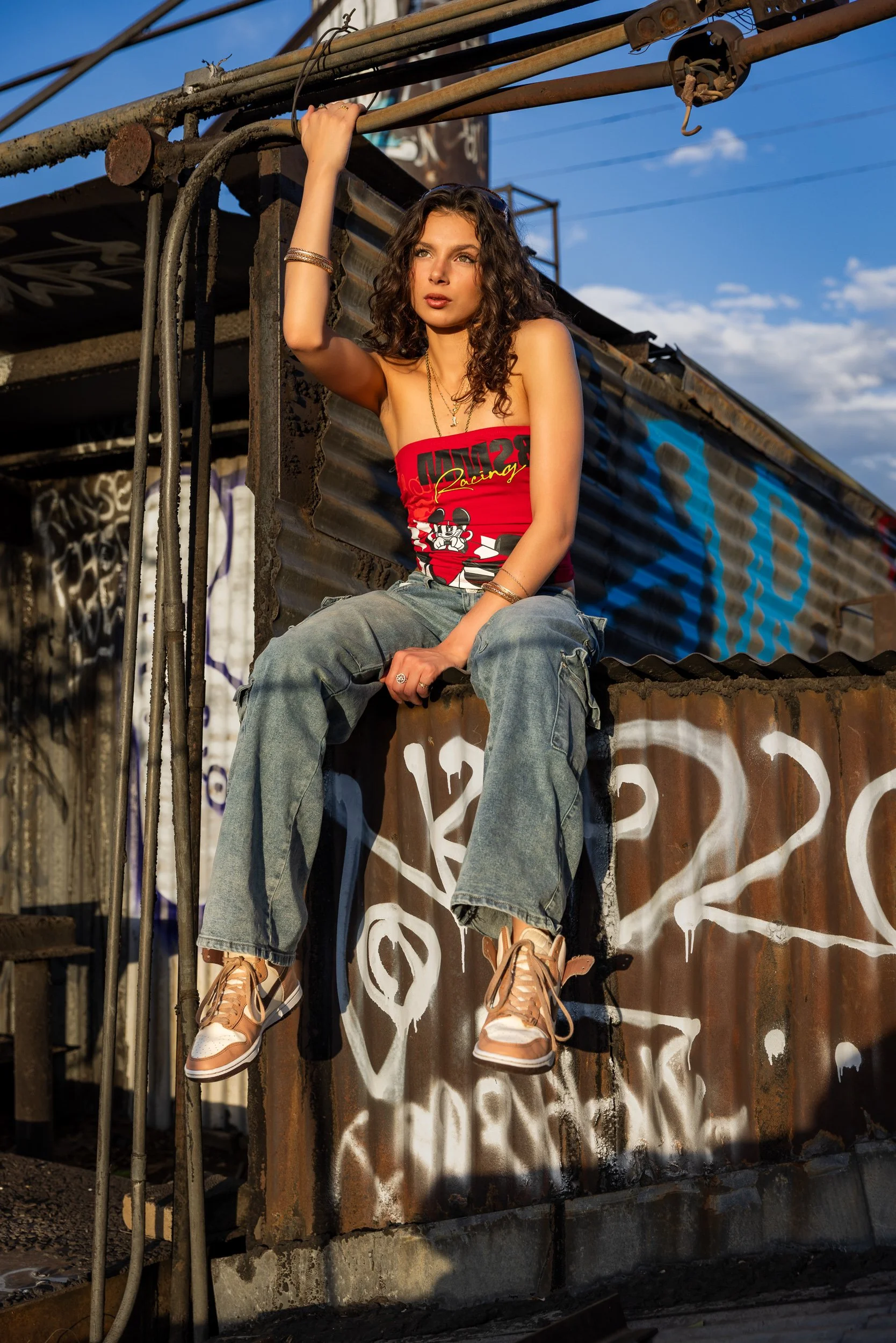 A young woman with curly brown hair wearing a red tank top with racing graphics and baggy jeans, sitting on a rusted metal structure in an urban setting during sunset. She has multiple jewelry pieces, including bracelets and necklaces, and is looking