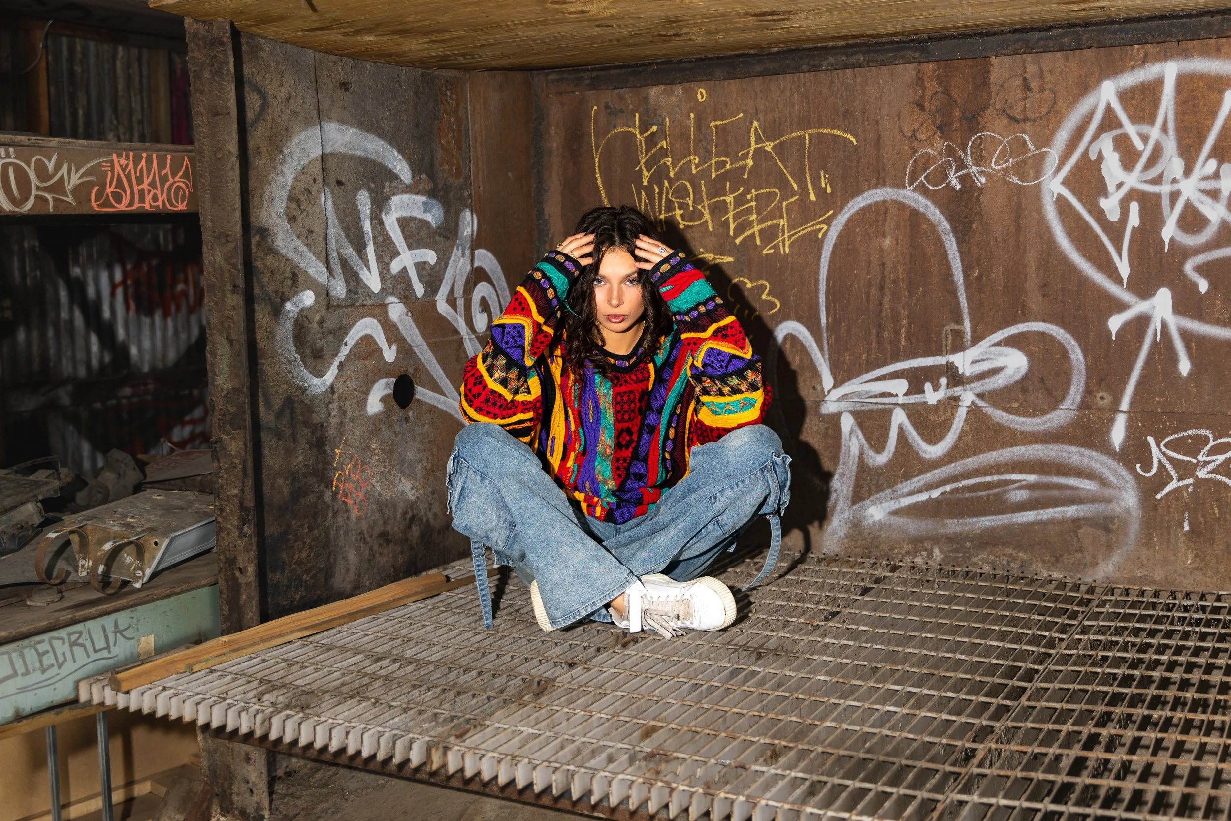 A young woman with curly hair wearing a colorful sweater and jeans, sitting cross-legged on a metal grate floor in an urban setting with graffiti-covered walls behind her.