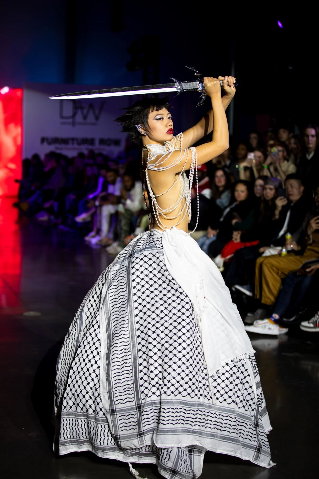 A woman in a dramatic black-and-white gown with layered pearl accessories holds a large sword above her head during a fashion show. The runway is filled with spectators and a backdrop featuring the text 'FURNITURE ROW'.
