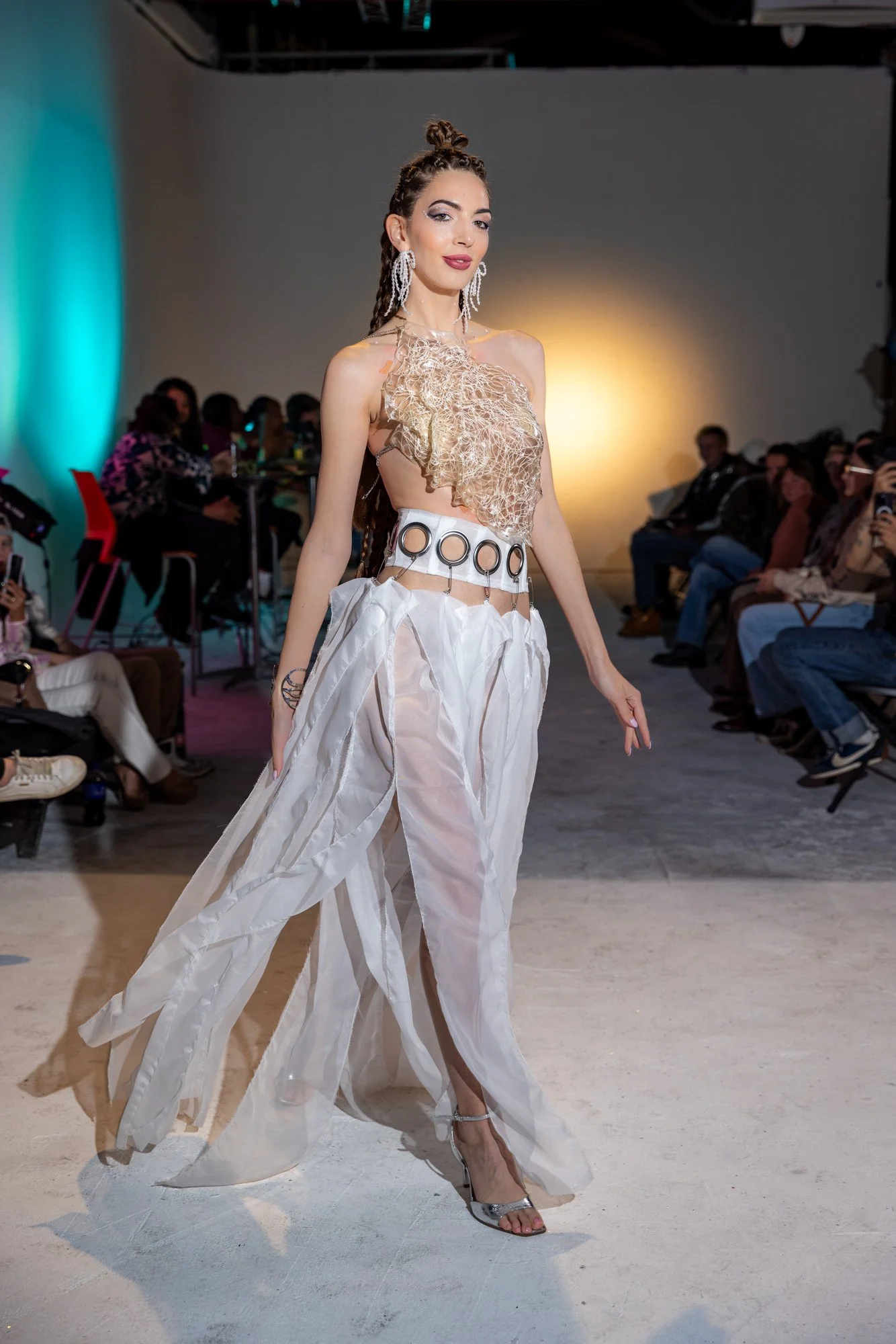 A fashion model walks down the runway at a fashion show, wearing a beige and white outfit with intricate details, large jewelry, and high heels, with spectators watching and taking photos in the background.