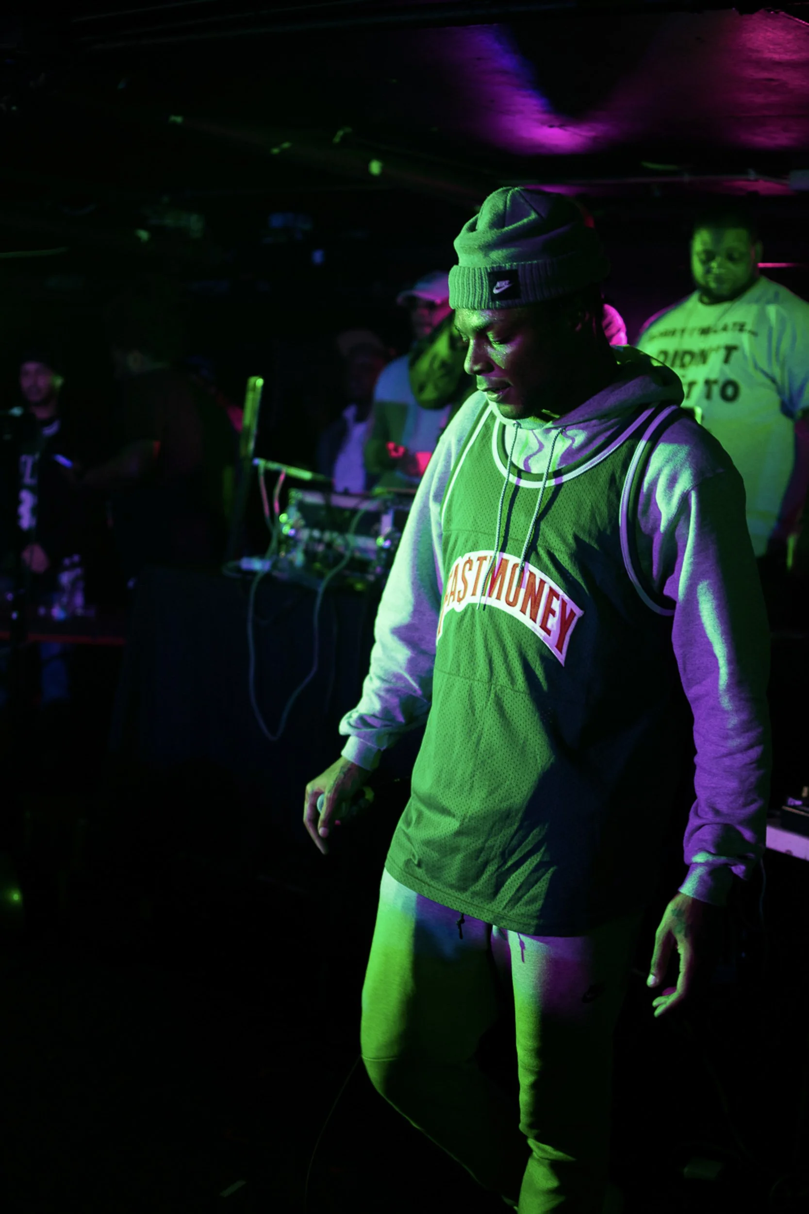 A young man in a beanie and sportswear DJing at a nightclub with colorful lighting, with other partygoers in the background.
