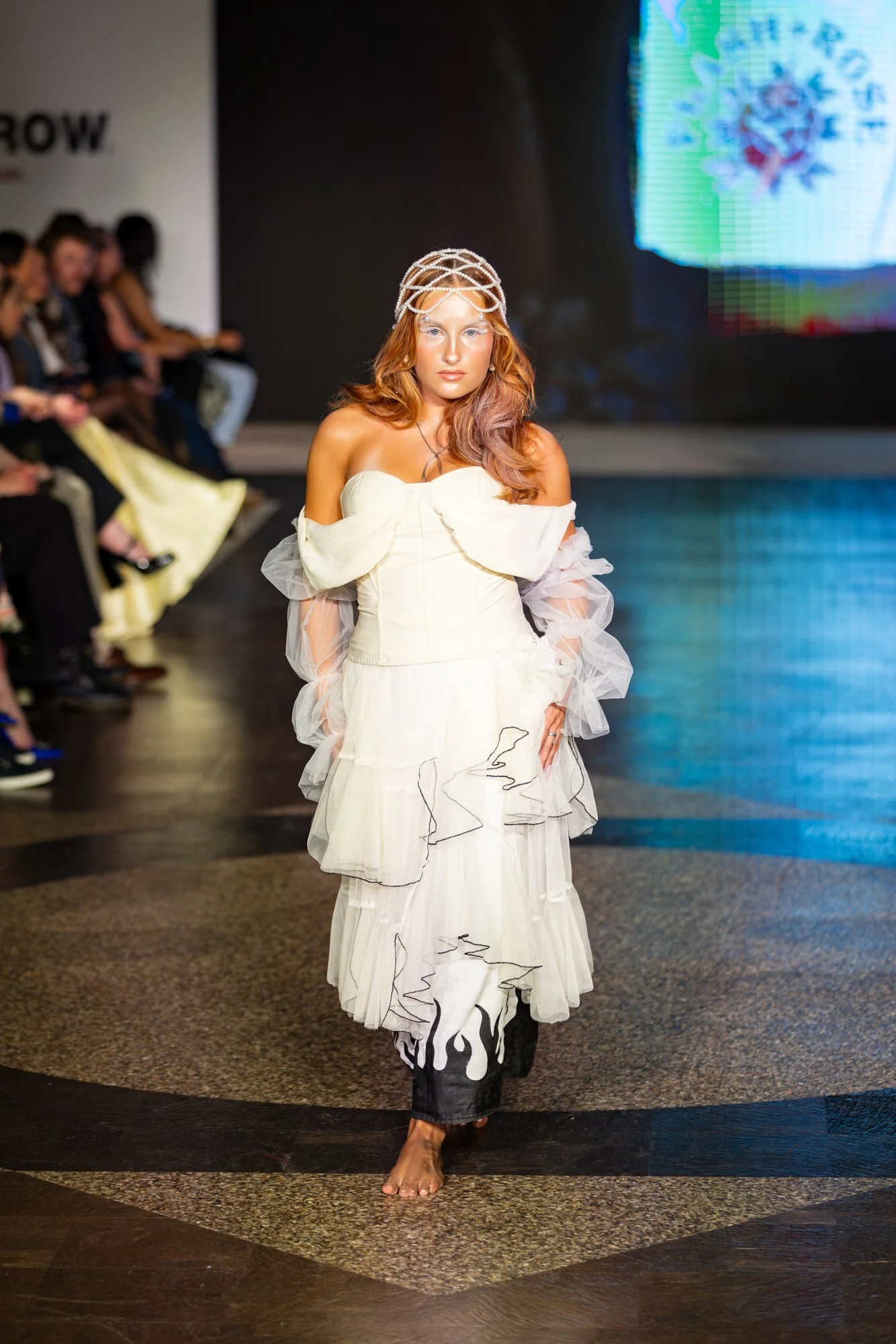 A woman walks barefoot on a runway during a fashion show, wearing a white off-shoulder dress with ruffled sleeves and an intricate crown. Audience members are seated along the runway in the background.