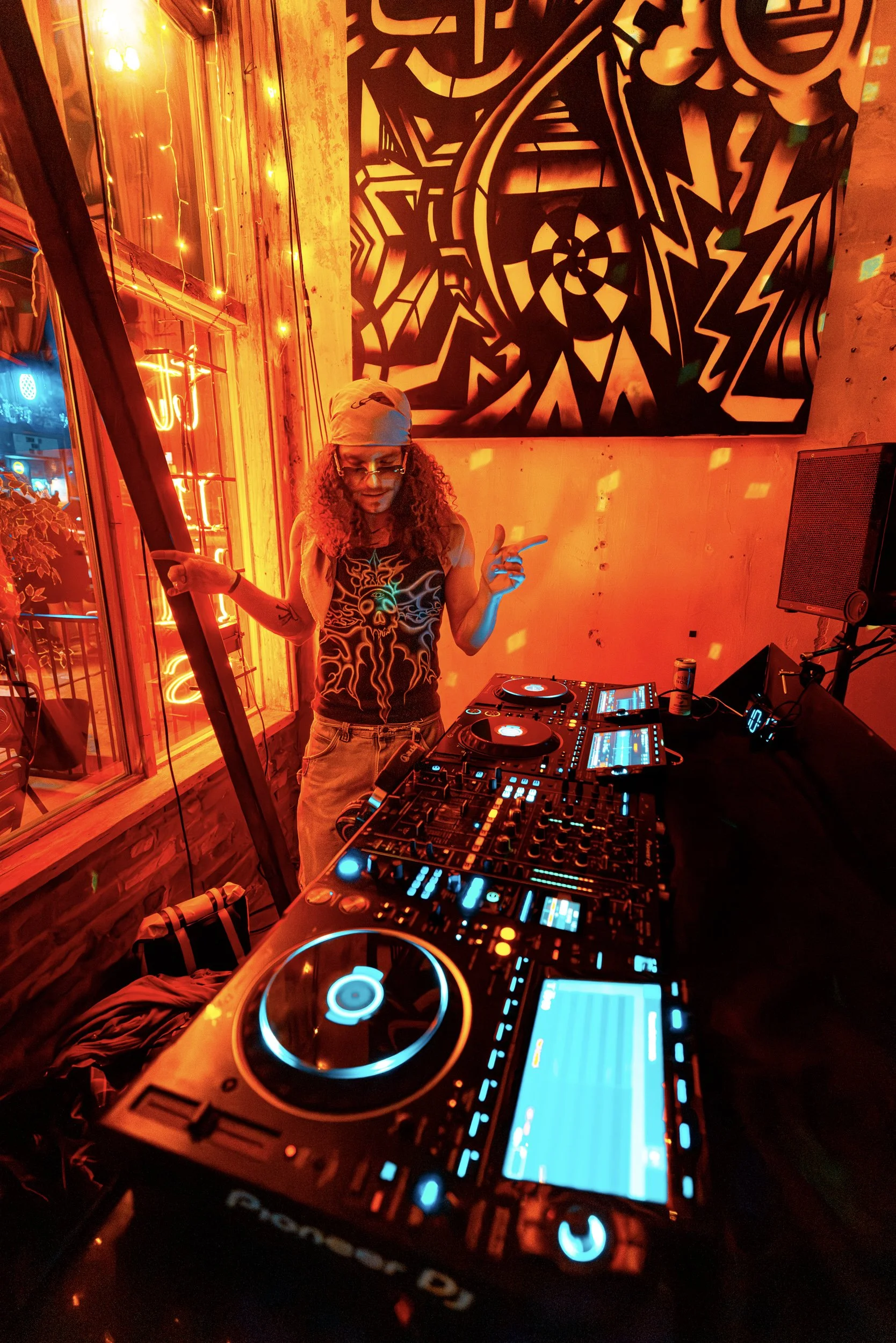 Person DJing at a club with illuminated DJ equipment and vibrant orange lighting, graffiti-style artwork on the wall, and decorative string lights outside the window.