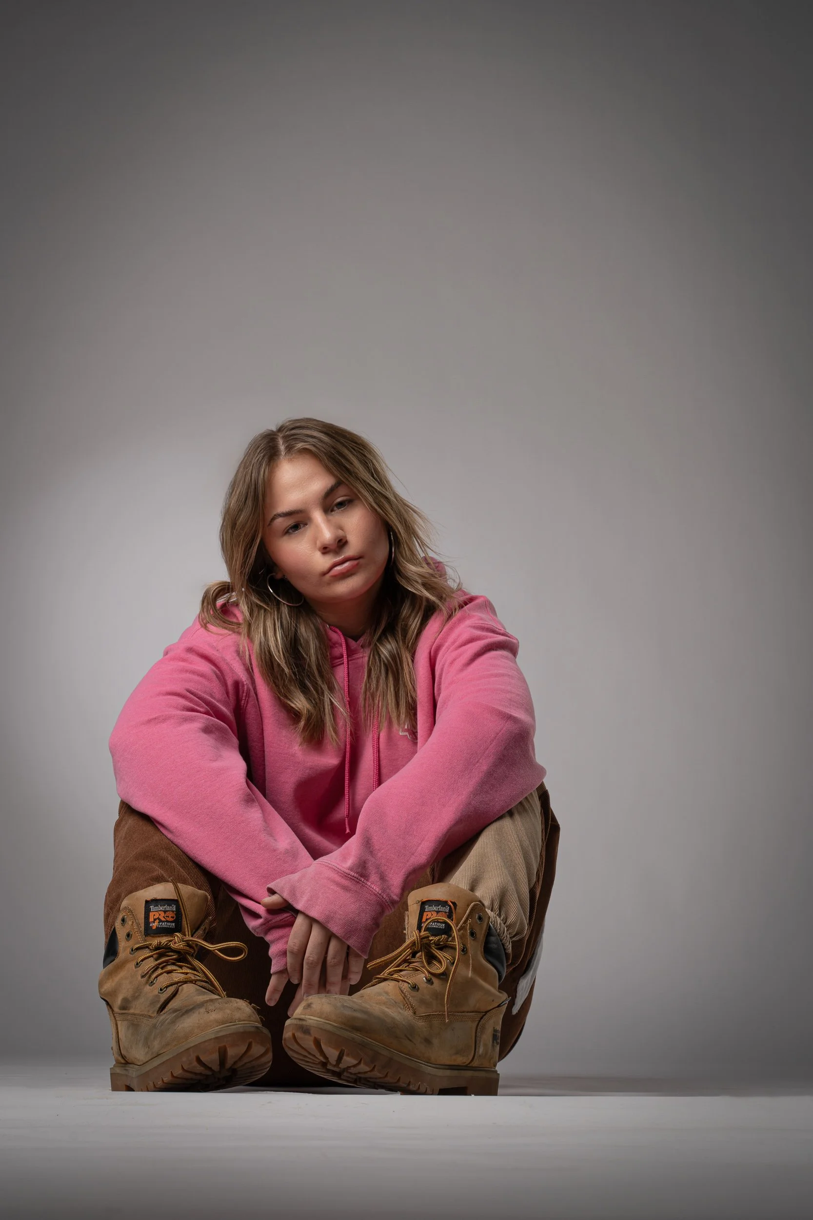 A young woman with light brown hair sitting cross-legged on the floor, wearing a pink hoodie, tan Timberland boots, and beige pants, against a plain gray background.