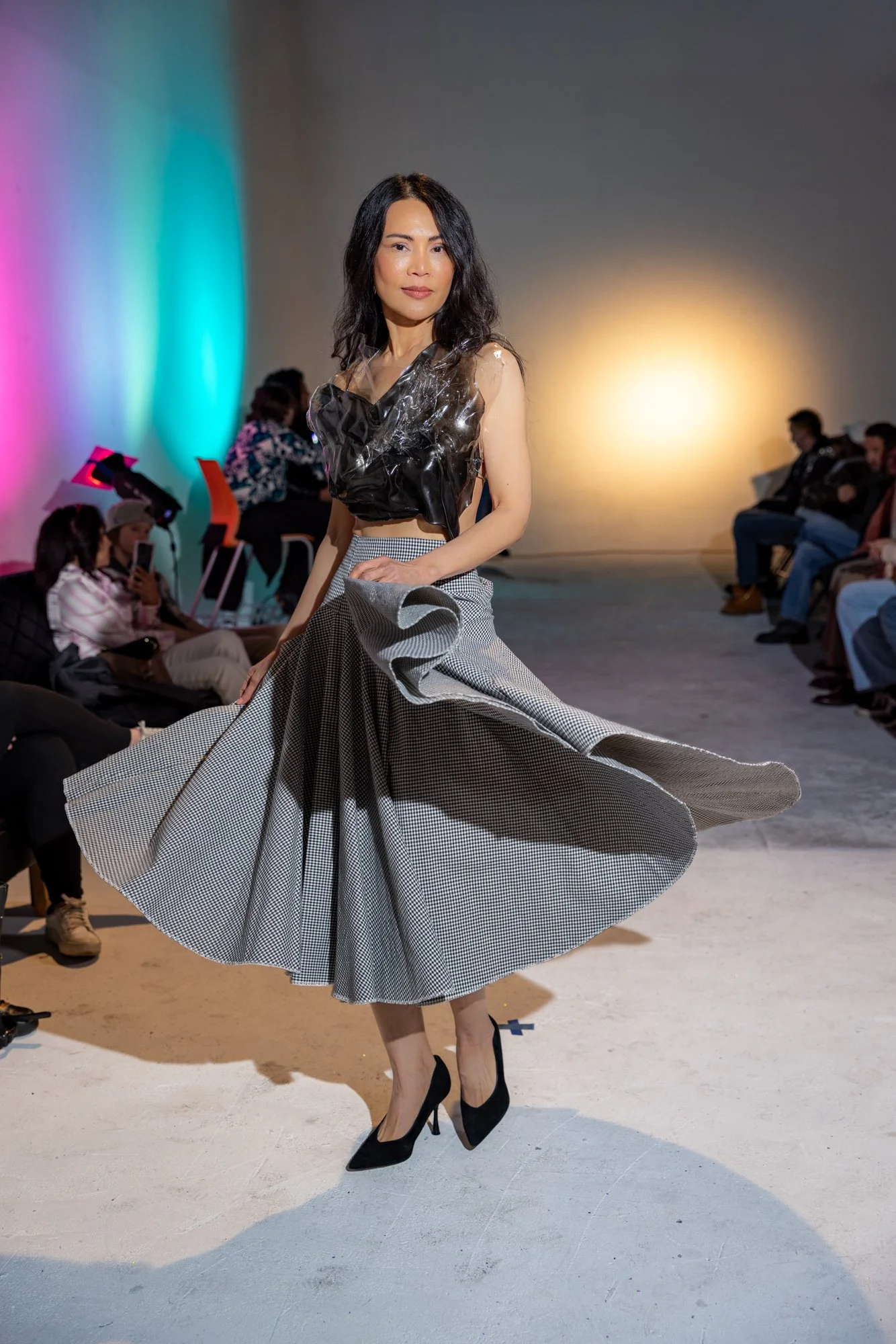 A fashion model is walking on a runway wearing a black top with shiny details and a big gray patterned skirt. She has long black hair and wears black high heels. The background shows an audience seated on either side of the runway, with colorful ligh