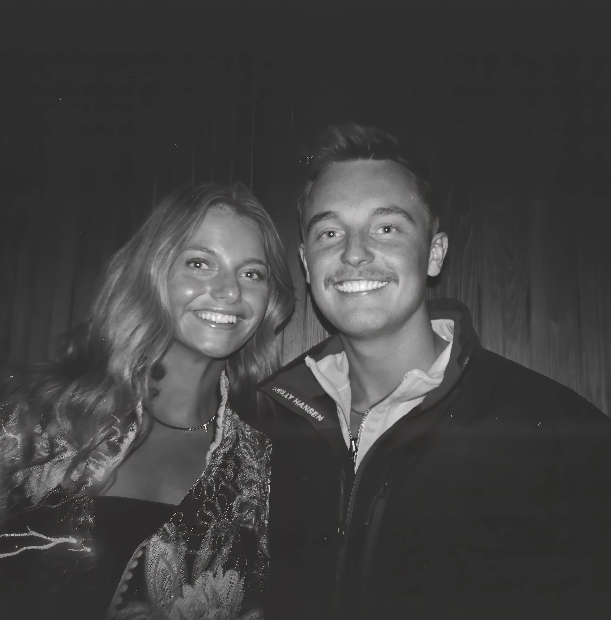 Black and white photo of a smiling woman with long hair and a smiling man with short hair, both dressed in formal attire, posing together against a dark background.