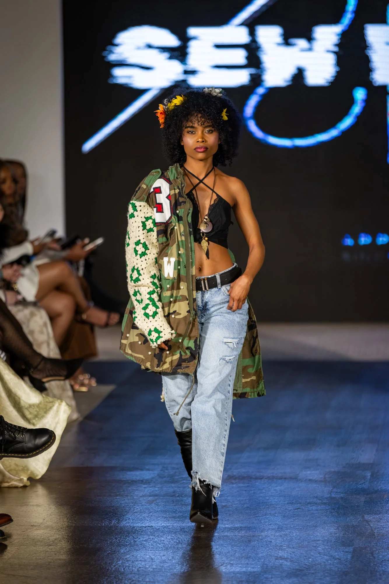 A female model walking down a runway wearing a black crop top, distressed jeans, black boots, and a camouflage jacket with patches and knit sleeve, accessorized with layered necklaces. She has curly hair decorated with colorful flowers and is walking