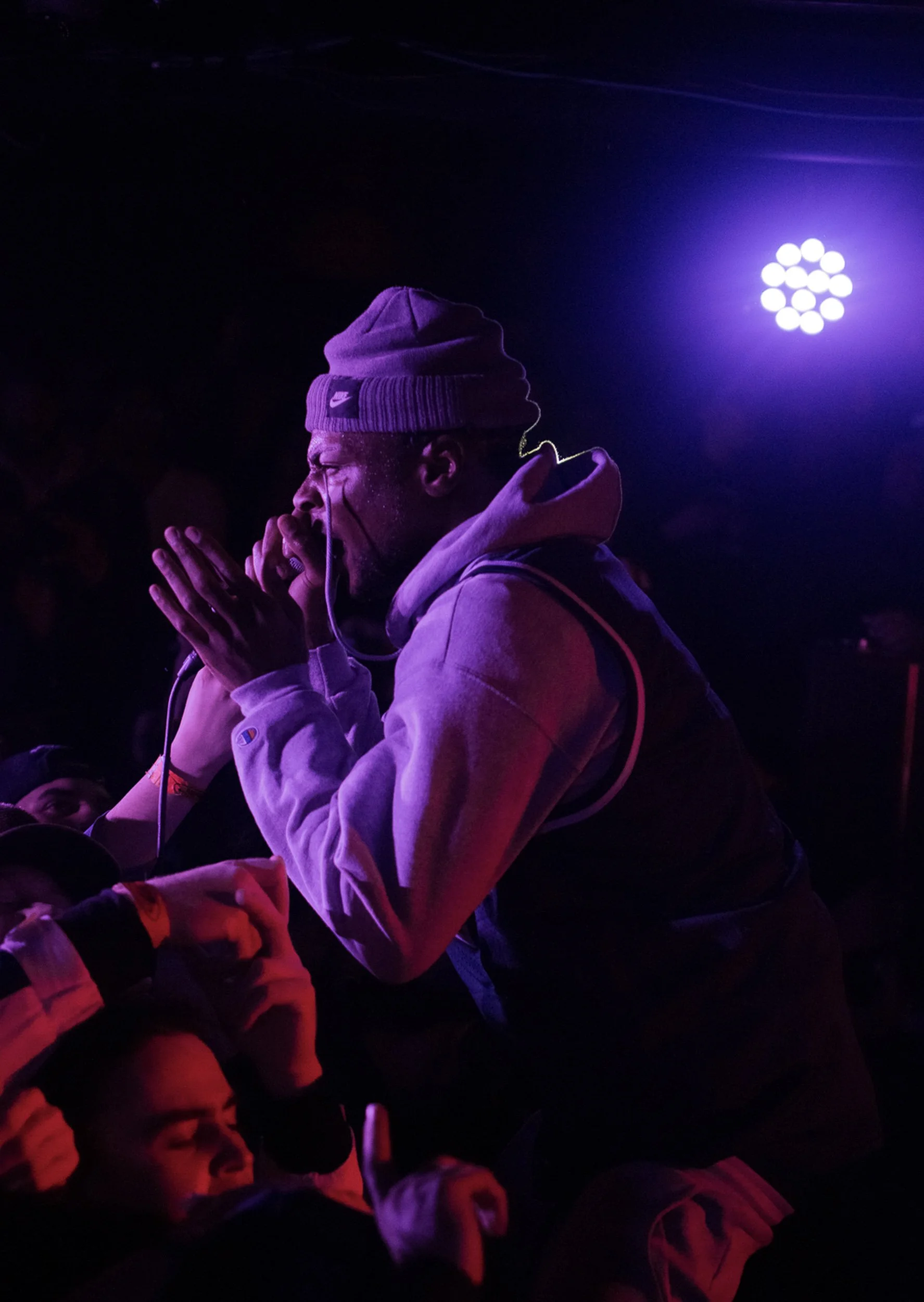 A person in a gray hoodie and beanie hat is praying or meditating with hands pressed together, surrounded by other people, with purple stage lighting in the background.