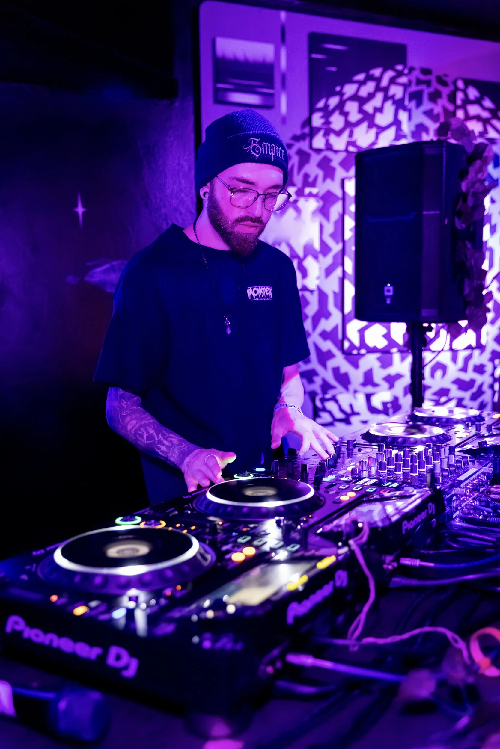 A DJ wearing glasses, a beanie, and a black t-shirt with 'Monster' written on it, performing with DJ equipment in a purple-lit setting with patterned walls and a speaker in the background.