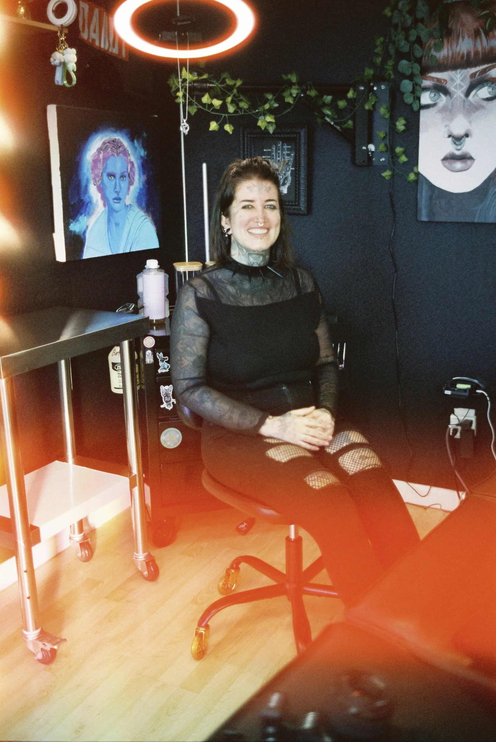 A woman sitting on a black rolling chair in a room with dark walls, colorful artwork on the walls, and a neon light hanging from the ceiling. She has a septum piercing, tattoos, and is smiling at the camera while wearing a sheer black top and fishnet