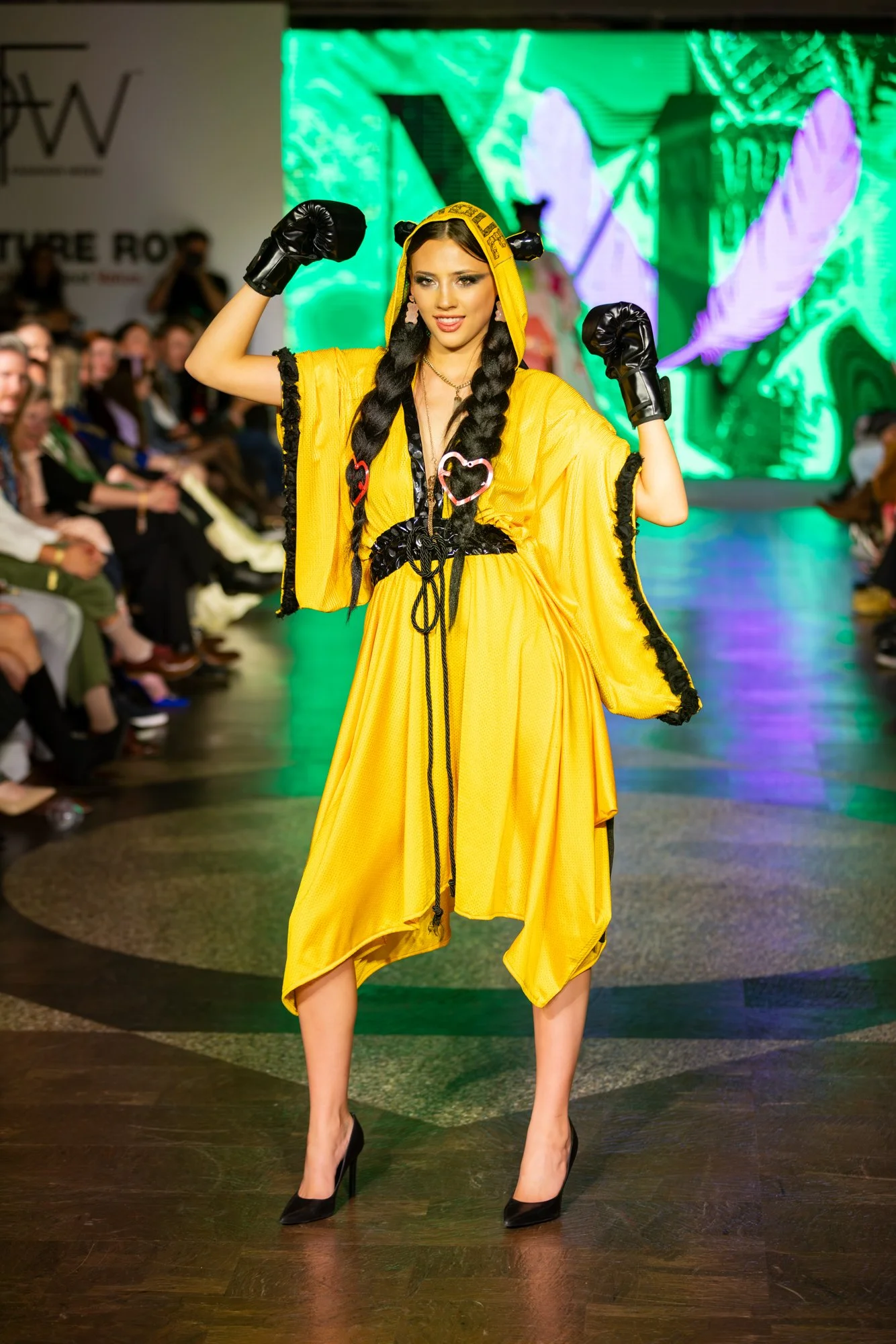 A woman with braided hair in a yellow dress and black high heels is posing with her arms flexed at a fashion runway show. She is wearing black gloves and has decorative heart-shaped accessories on her dress. The audience is seated on either side, and