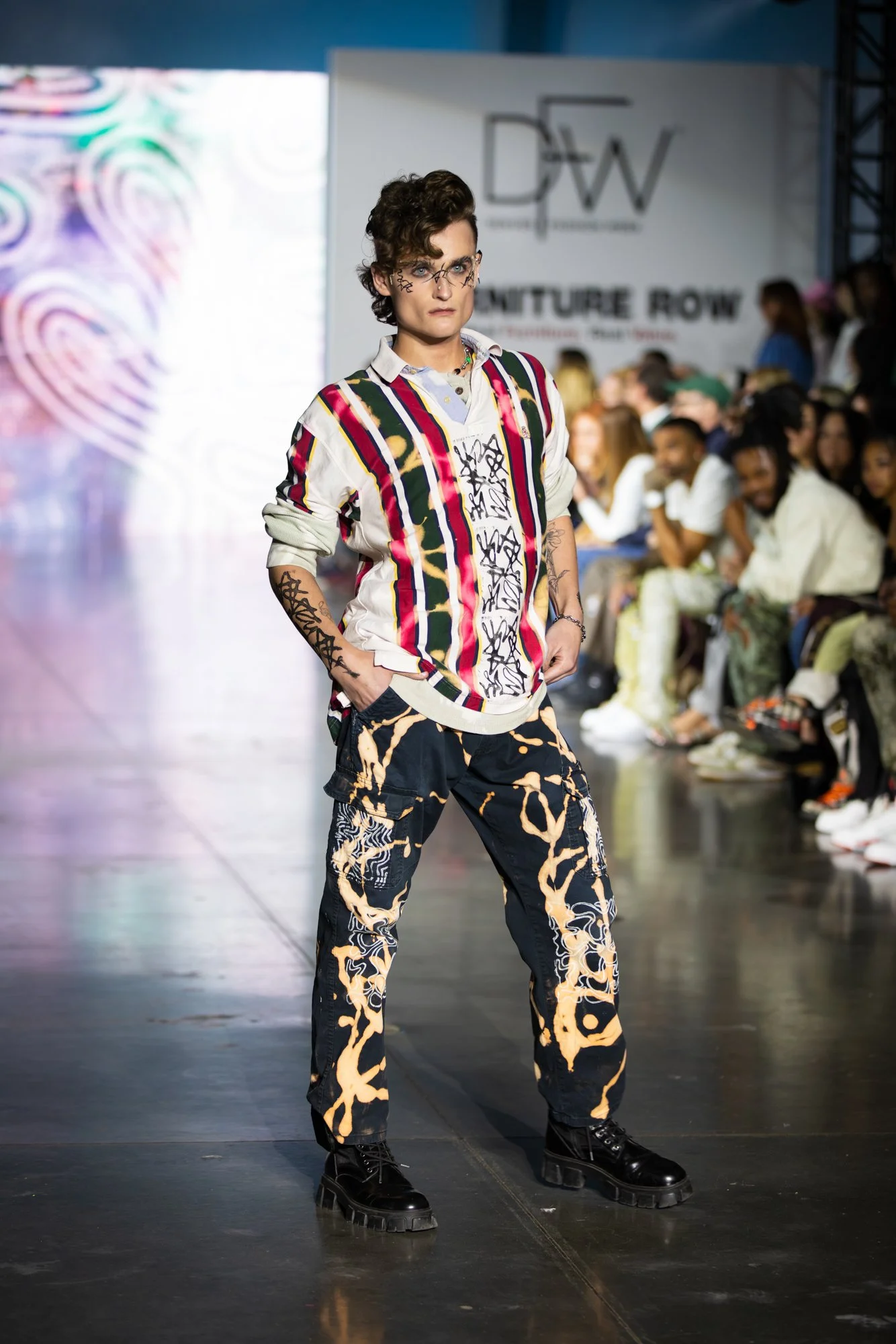 A male model walking on a runway at a fashion show, wearing a colorful striped shirt with graphic patterns, patterned pants, and chunky black boots, with an audience and a 'D&W' sign in the background.
