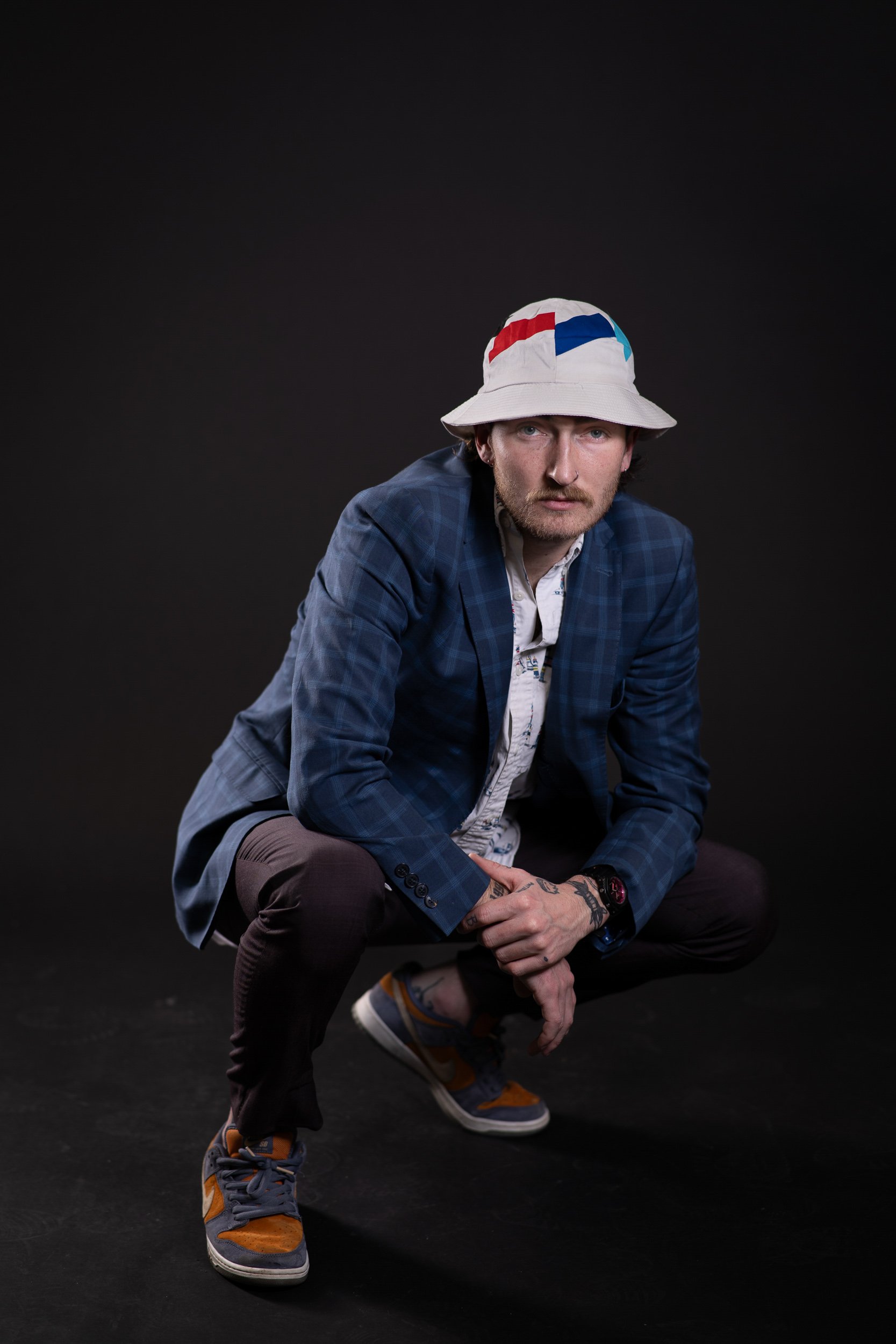 A man crouching against a black background, wearing a white bucket hat with colorful red, blue, and teal patterns, a blue plaid blazer, a white patterned shirt, dark pants, and gray and orange sneakers. He has a mustache, beard, tattoos on his hands,